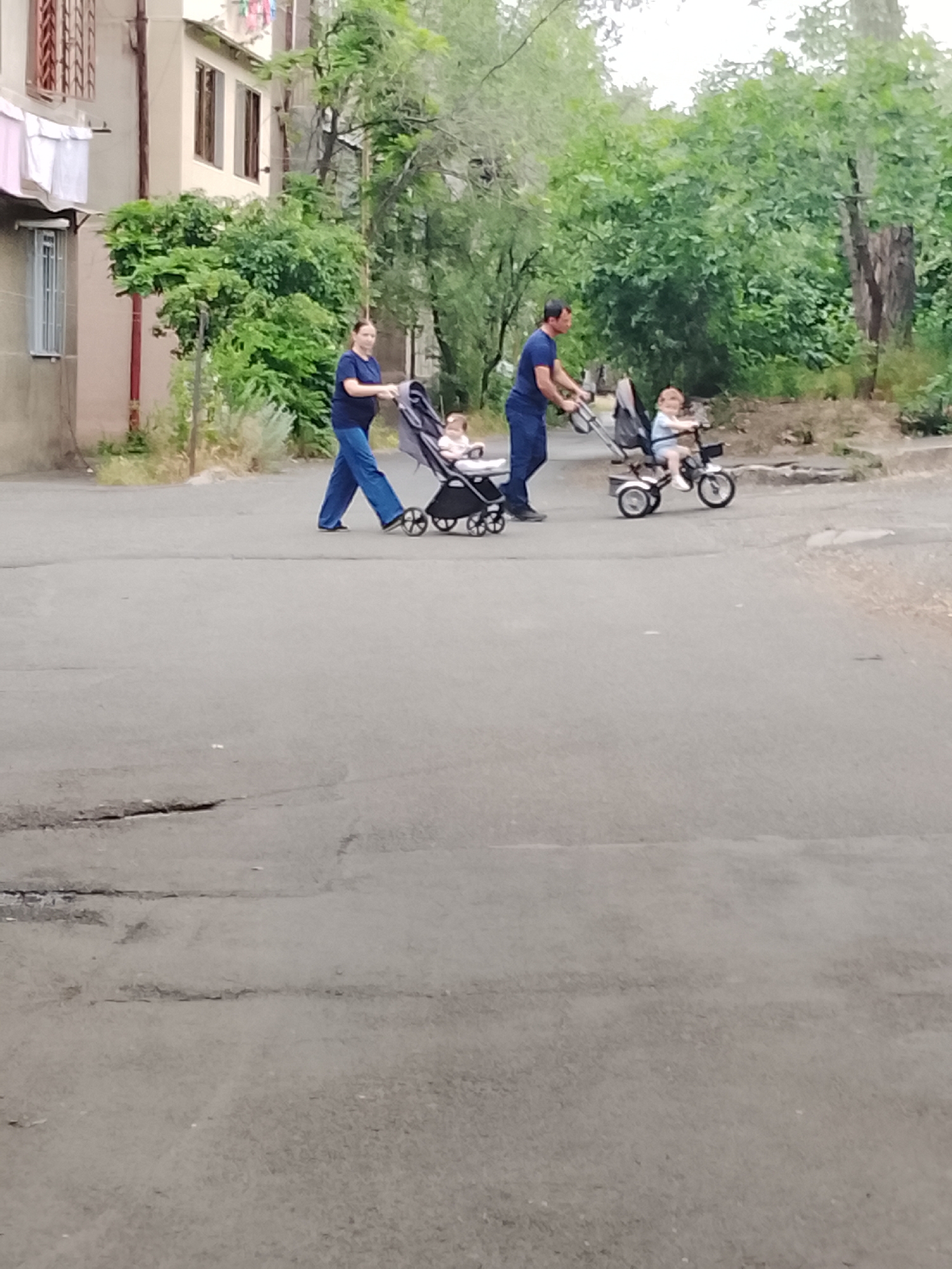 In an outdoor, urban environment with a cracked asphalt road, a woman in blue pants and a dark shirt pushes a stroller with a baby. To her right and slightly behind her, a man in a blue shirt and dark pants pushes a second stroller, which appears to be a tricycle, with a child riding in it. Lush green trees and a building are visible in the background.