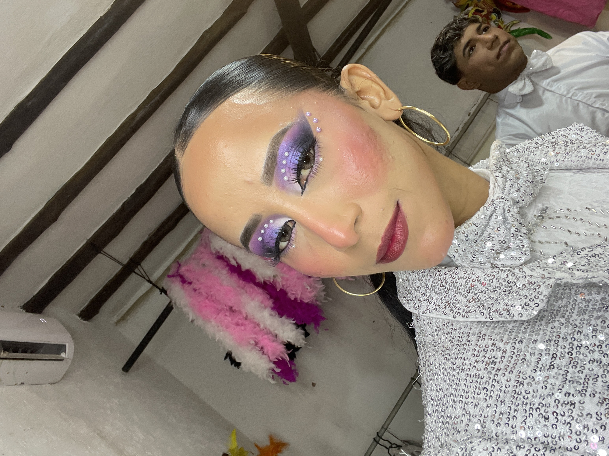 In an indoor setting, possibly a dressing room, a woman with dramatic purple and white eye makeup, adorned with small pearl-like accents, poses for a selfie. She wears a heavily sequined white jacket with a prominent, sparkling bow tie. A man in a white shirt and bow tie stands in the background, looking away from the camera. Behind the woman, a cascade of pink and white feathers hangs on the wall, suggesting a festive or theatrical context. The room appears to have exposed wooden beams on the ceiling and a white air conditioning unit. The overall impression is one of preparation or backstage activity for a performance. The city of Porlamar, Venezuela, is not explicitly depicted, nor are there any identifiable landmarks that would confirm its location.