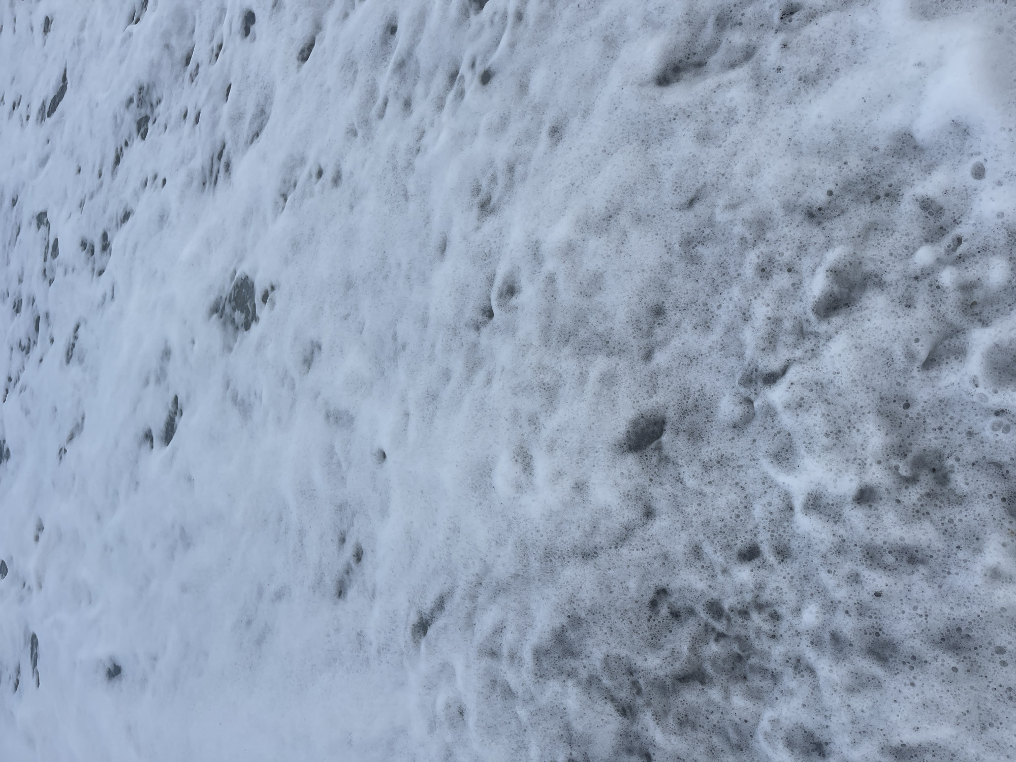 The image shows an area covered in thick, white foam. The foam has a bubbly texture and appears to be partially obscuring a dark surface underneath. There are variations in the density and thickness of the foam across the frame. The location is Unknown, Unknown. No people or objects are visible other than the foam and underlying surface.