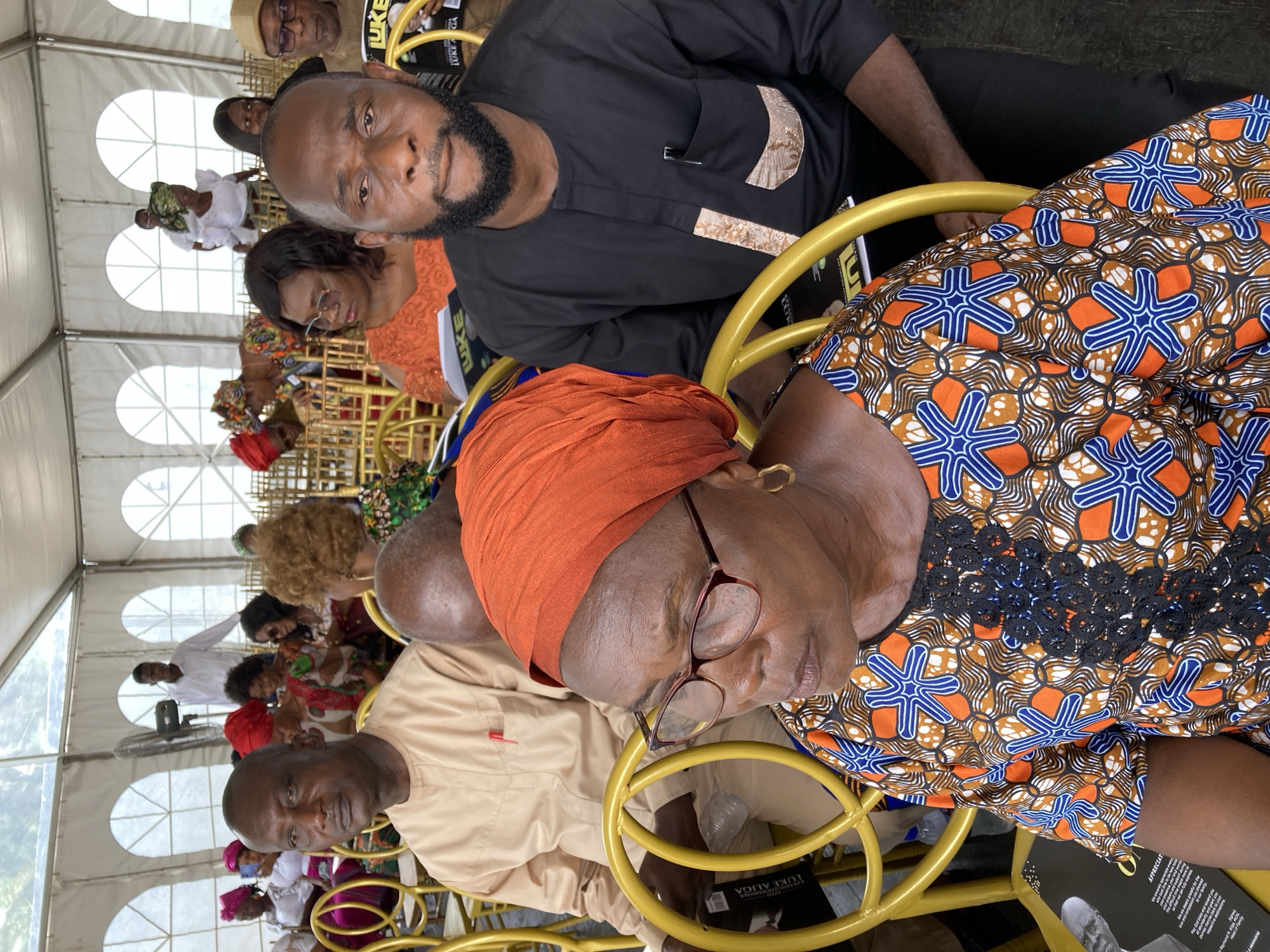 A medium, portrait-style shot shows a group of people sitting on gold-colored metal chairs inside a tent. The location is identified as Nnenu, Nigeria.
Several individuals of varying ages and dressed in a range of attire are visible. In the foreground, a woman wearing an orange headscarf, glasses, and a patterned orange and blue dress is seated. Next to her, another person in a tan shirt is visible. Behind them, a man with a beard wears a black shirt with decorative accents. Further in the background, more people can be seen sitting, with some wearing traditional Nigerian clothing.
The environment is defined by the white tent, which is made of translucent material allowing for soft light to fill the space. The chairs are simple in design and seem to be the seating arrangement for the event. A book or program, visible on the seat, might be the order of service for a burial service of IZEAGUORIANAMMA LUKE ALIGWE. Overall, the scene conveys a sense of community and gathering.