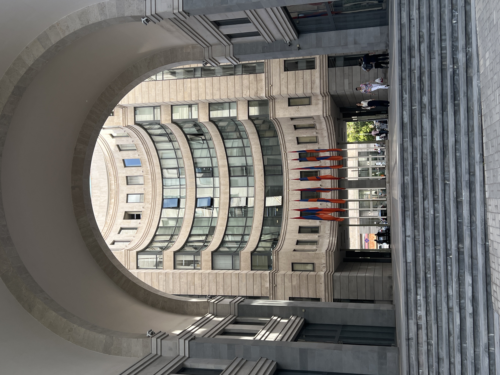 This image captures a grand architectural scene in Yerevan, Armenia, presented in bright daylight. The composition is framed by a massive, smooth, light-colored archway on the left, made of large stone blocks, which curves inward to reveal the interior space.
Beyond the archway, a prominent multi-story building with a striking curved facade dominates the view. Its exterior is clad in light beige stone, characterized by numerous dark-framed windows, many of which are also curved to follow the building's form. Several windows are ajar. The building's design features horizontal lines and subtle variations in depth, adding texture to its surface.
On the right, a substantial staircase or stepped platform of grey stone or concrete extends into the midground, marked by strong vertical grooves. Several people are seen walking across this public space. One individual in a white and patterned top, light pants, and carrying a bag, is visible, alongside others in darker clothing. In the distant background, through an opening, more urban structures and foliage are discernable, including what appears to be a red stop sign.
In front of the curved building, a line of flagpoles proudly displays the flags of Armenia, with their characteristic red, blue, and orange horizontal stripes. The clear, bright sunlight and distinct shadows indicate it is daytime, likely midday or early afternoon. The scene portrays an active urban environment with people going about their daily routines amidst impressive architecture.