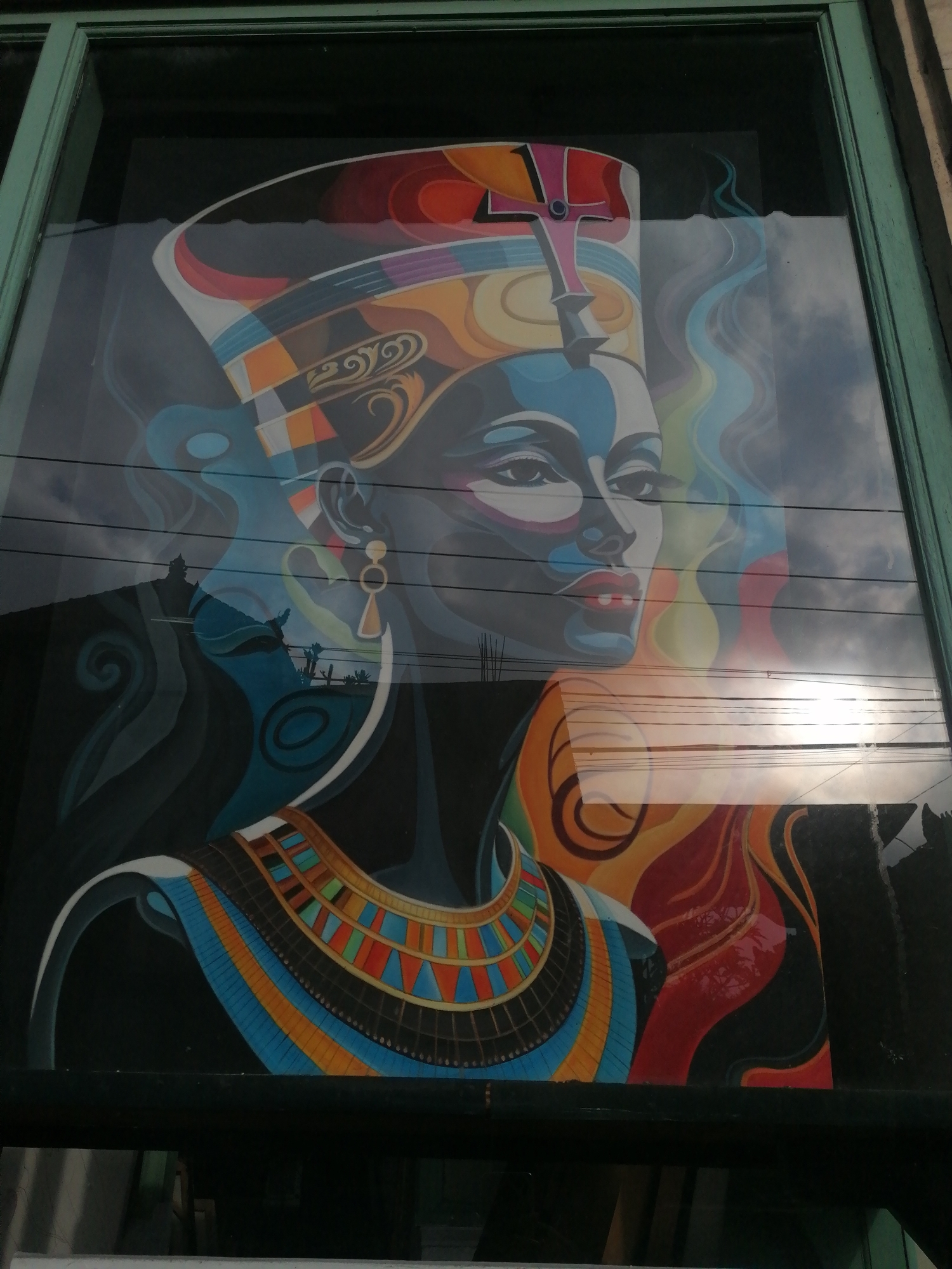 An image shows a colorful mural on a window of a building in Ubud District, Indonesia. The mural depicts an Egyptian-style portrait of a woman wearing a traditional headdress and elaborate jewelry. The woman's face is rendered in shades of blue, black, and white, with red lips and dark eyes. The headdress features vibrant colors including orange, yellow, purple, and blue. She is also wearing a large, ornate collar with geometric patterns in similar bright colors.
The mural is seen through a glass window, which reflects the sky and surrounding environment. The sky appears cloudy with sunlight breaking through, creating bright reflections on the glass. Several power lines are visible, crossing horizontally across the image, partially obscuring the mural and its reflection. The building has a dark green frame around the window. The overall scene is a juxtaposition of the traditional art depicted in the mural and the contemporary urban elements of the reflections and power lines.