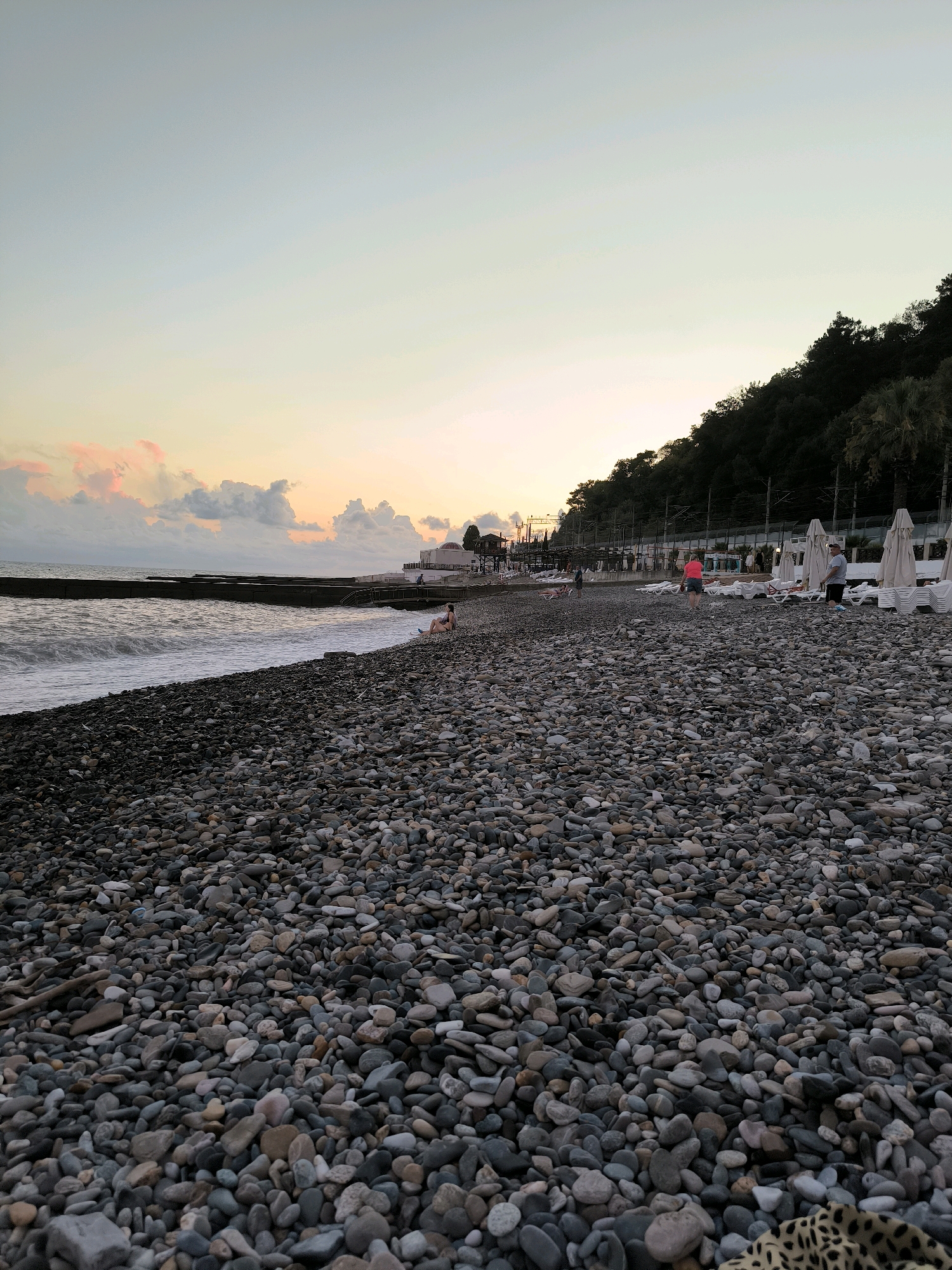 This is a daytime, eye-level, full shot of a pebble beach on the coast of Sochi, Russia. The sky is a gradient of pale blue to a very light orange, with scattered pink and white clouds. The sea is a muted grey-blue with gentle waves. In the foreground, the beach is covered in smooth, grey, and tan pebbles.
Along the waterline, there is a concrete breakwater. In the middle ground, several people are visible on the beach and near the water. A person is sitting on the pebbles, and two people are walking. Further back, rows of empty white lounge chairs and umbrellas are set up. Behind the lounge chairs, there is a dark green, tree-covered hillside that slopes upwards. A few palm trees are visible on the hillside. In the distance, a white building with a red-tiled roof can be seen. Power lines and poles run along the base of the hillside. The overall scene is tranquil, with a few people enjoying the beach at dusk.
