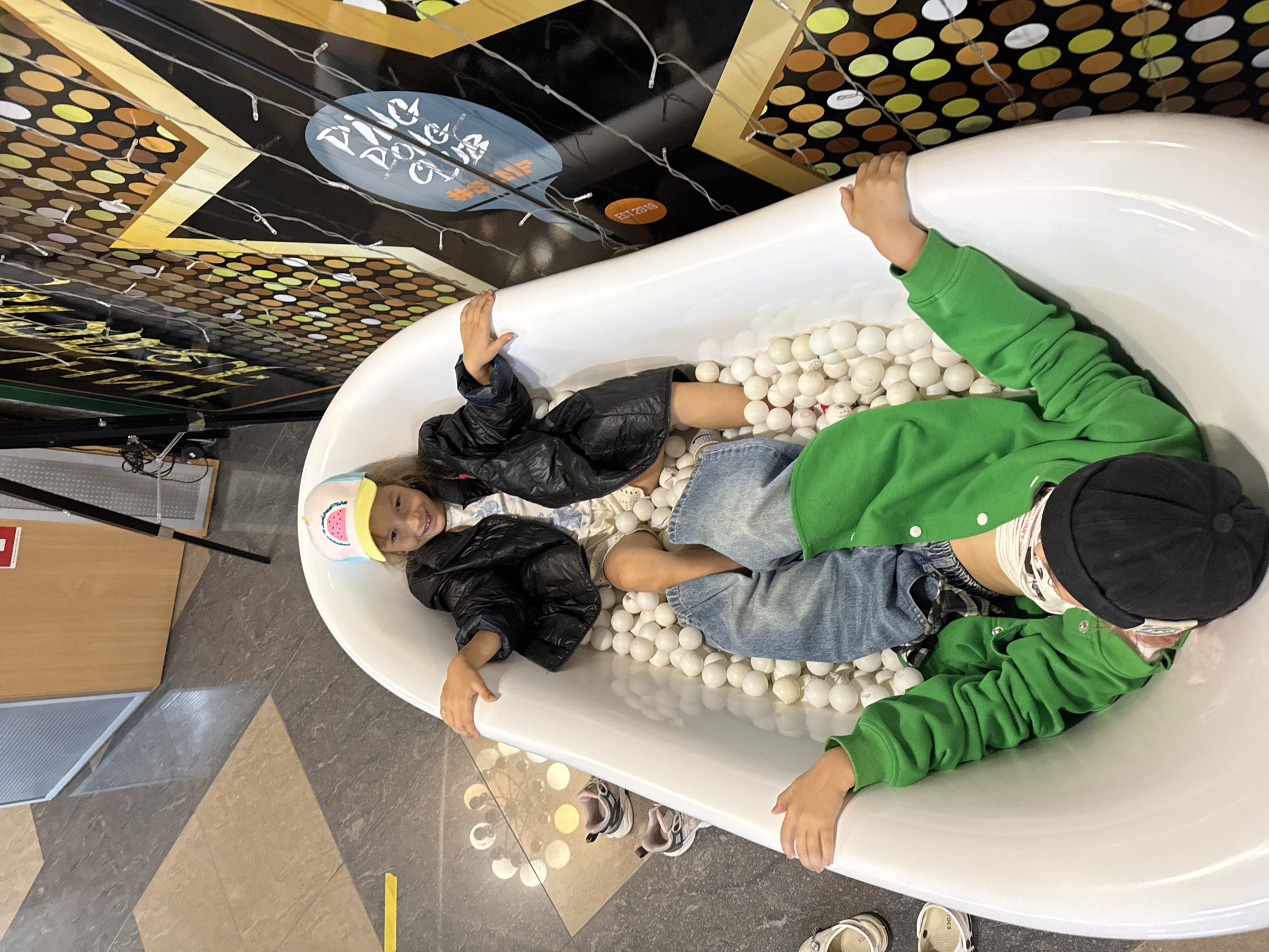 Two children are in a white bathtub filled with white balls. One child, wearing a black puffer jacket and a hat with a watermelon design, is sitting up and smiling. The other child, wearing a green hoodie and a black cap, is lying upside down with their legs in the air. The bathtub is on a tiled floor, and a decorated wall with circular patterns and lights is visible in the background.