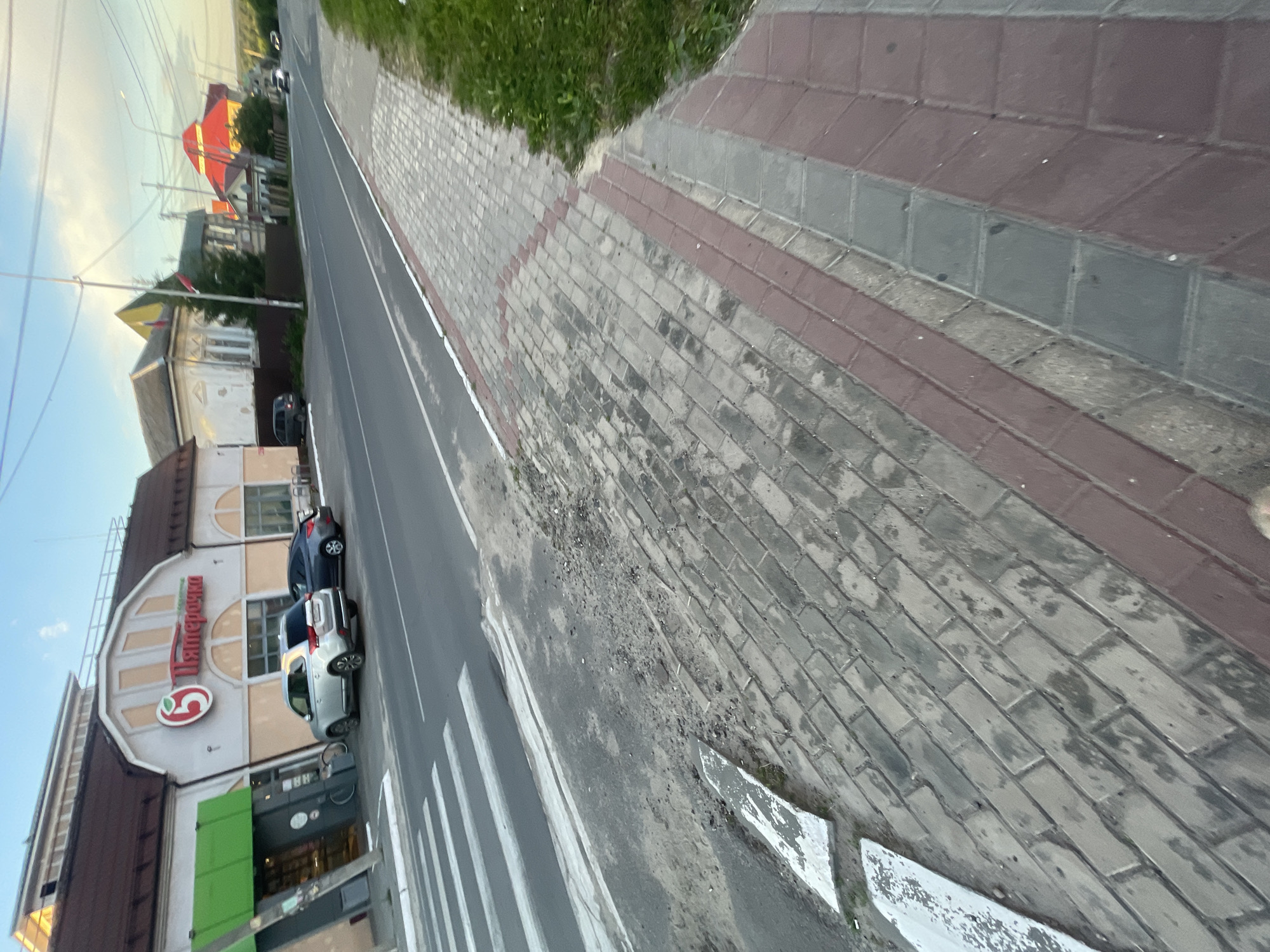 The image depicts a street scene in the city of Krasnye Baki, Russia. In the foreground, a pedestrian sidewalk made of gray and reddish-brown pavers is visible, with grass bordering the right side. A crosswalk with white stripes is on the left, adjacent to the sidewalk. Beyond the crosswalk, a street runs parallel to the sidewalk, with two cars parked on the right side of the street. One car is silver, and the other is dark gray. Further down the street, a commercial building with a brown roof and light-colored facade is visible, featuring a prominent red circular logo and Russian text, likely a store. The building has several windows and an entrance with a green awning. Behind the commercial building, residential houses with pitched roofs can be seen, some with red roofing material. The sky is partly cloudy with a hint of blue. The overall scene suggests a typical daytime view of a street in a Russian town or city.
