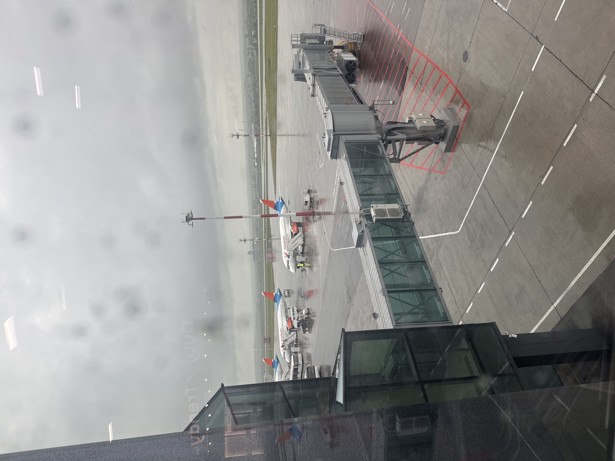 The image depicts an airport tarmac on a rainy day. Three airplanes are visible, each with an orange and blue tail. One jet bridge is connected to an airplane. The scene includes airport vehicles and staff around the planes. The image is taken from inside an airport terminal, looking out through a window. The location is Unknown, Unknown.