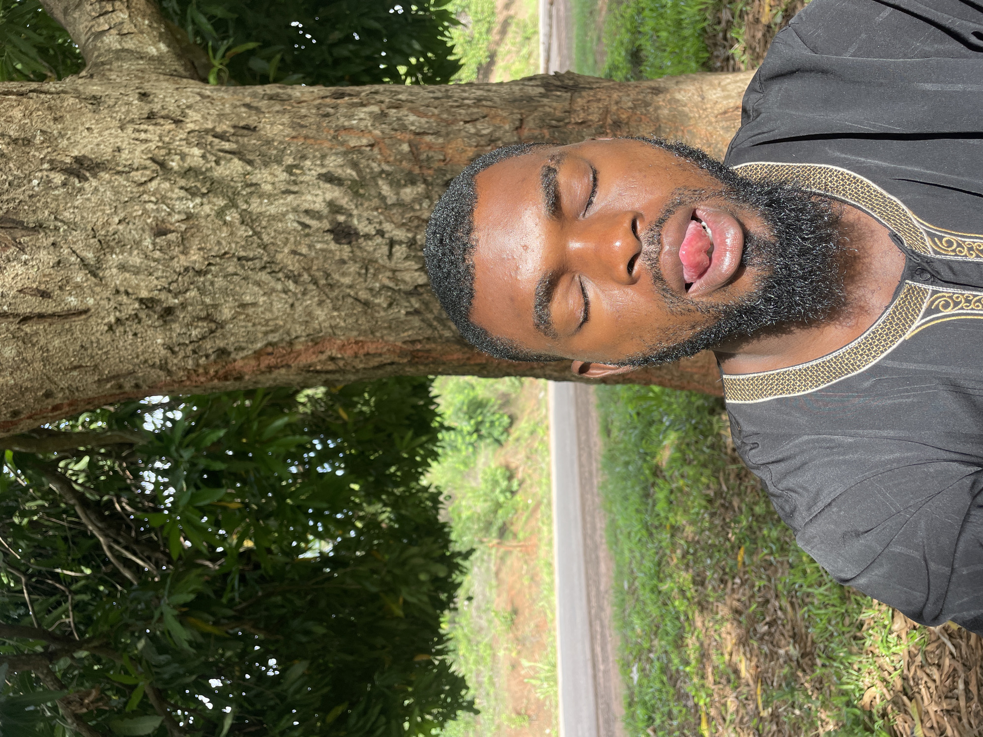 A man with dark skin and a black beard stands in front of a tree in Ogbe, Nigeria. He is wearing a black shirt with gold embroidery around the collar. His eyes are closed, and his mouth is open, with his tongue sticking out. In the background, there is a road and green vegetation.