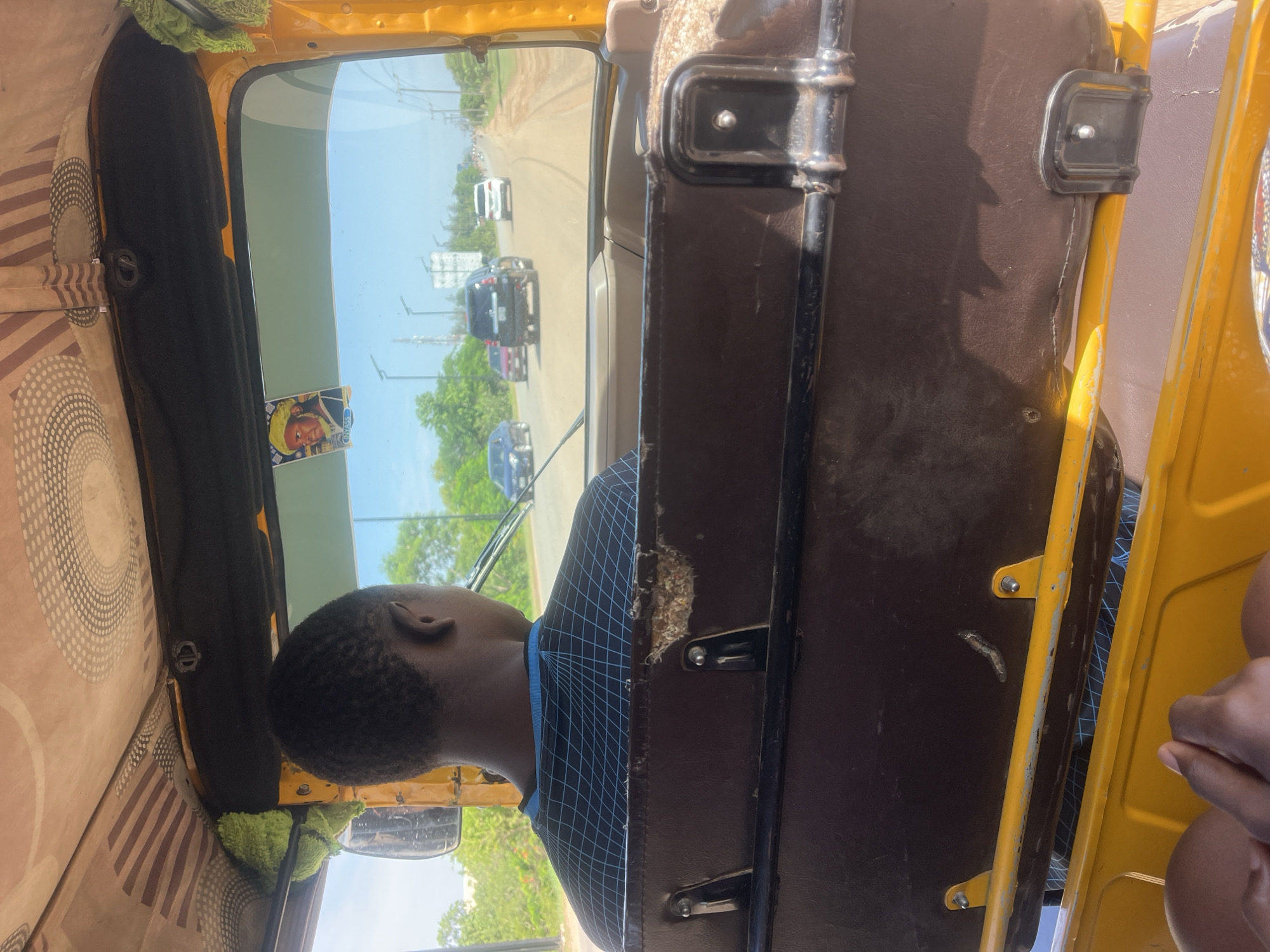 The image captures a rear interior view from a yellow auto-rickshaw in Galadimawa Village, Nigeria, looking forward onto a road under clear daylight. In the foreground, the back of a driver's head and shoulders are visible; the individual has dark skin, short dark hair, and wears a black shirt with a blue grid pattern. Behind the driver, a worn brown seat with visible tearing and exposed material at the top is present, secured by metal bolts. Below the seat, yellow structural elements of the vehicle are apparent, with dark-skinned hands briefly visible at the bottom of the frame, likely belonging to the person taking the photograph.
The interior ceiling features beige fabric lining with brown striped and circular dotted patterns. A small green fluffy object hangs from the left side of the ceiling near a rearview mirror. Affixed to the upper part of the front windshield is a sticker depicting a person in a yellow turban, with the name "NYASS" written below the image.
Beyond the windshield, a paved road extends into the distance under a bright blue sky. Several vehicles are visible on the road: a black SUV, a white SUV, a red car, and a blue car. Green trees and vegetation line both sides of the road, and utility poles or streetlights are visible in the mid-ground. A billboard structure is also discernible on the left side of the road in the distance. The scene depicts travel on a main thoroughfare.