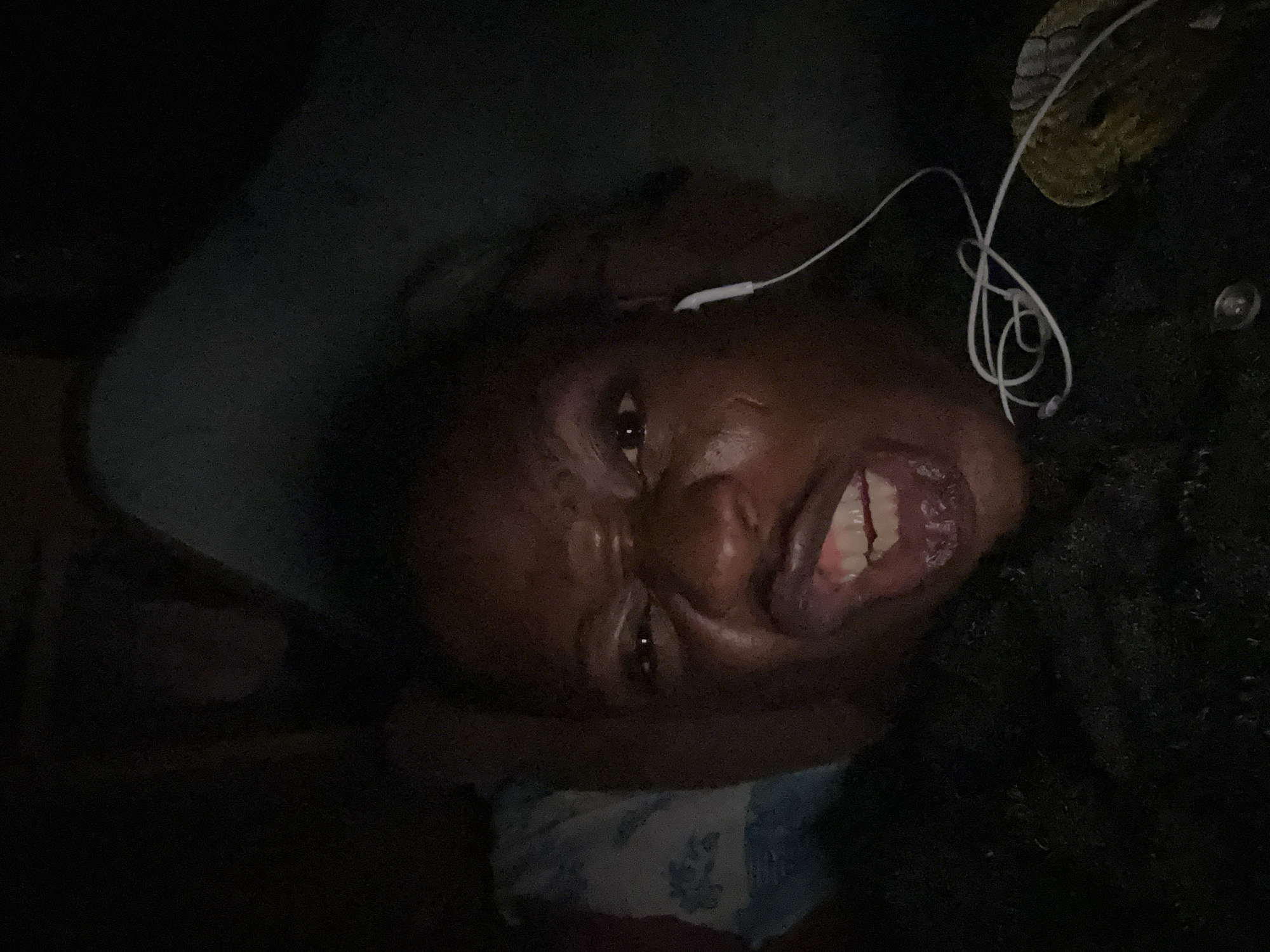 A dark, close-up, and somewhat distorted photograph depicts a person lying down, possibly on a bed or pillow. The person appears to be using wired earphones, one of which is visible in their ear. A patterned pillow or fabric is visible beneath the person's head. The environment seems dimly lit. The location and city are unknown.