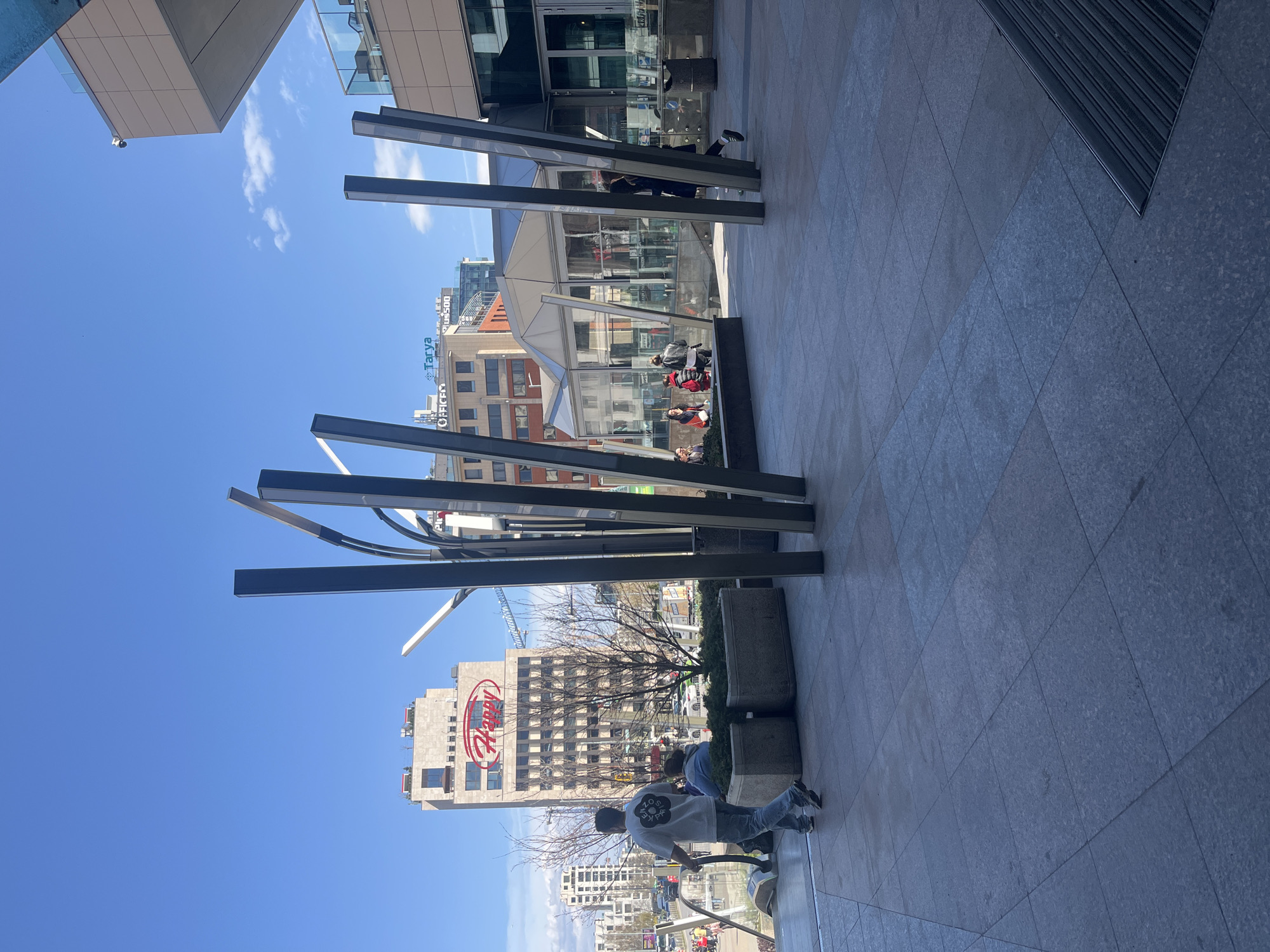 The image shows a public space in Sofia, Bulgaria, featuring modern architecture and a group of people. In the foreground, there are several dark, vertical metal structures arranged in an abstract design, partially obscuring the view. A person in a light gray t-shirt is visible in the lower right part of the picture. Behind the metal structures, a modern glass-enclosed structure houses people sitting and conversing. Beyond that, several buildings can be seen, including one with the "Happy" logo prominently displayed. Other buildings include the "Tarya" brand. The sky is blue with some clouds. The ground is paved with large, gray stone tiles.