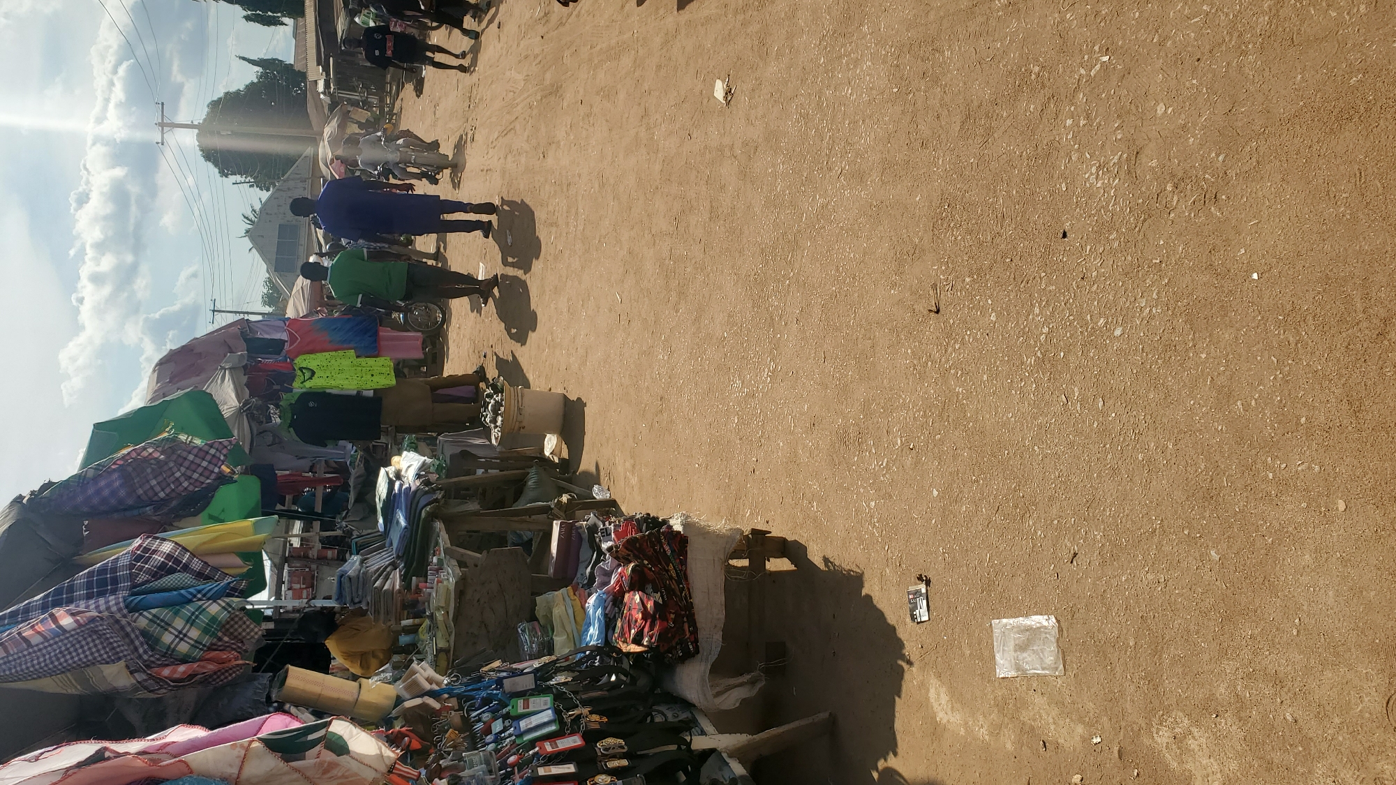 The image depicts an outdoor market scene in Jalingo, Nigeria, under a bright, sunny sky with scattered clouds. Stalls lined with colorful fabrics and clothing are visible on the left side of the frame, with various items displayed for sale. Several individuals are walking through the market area, which appears to be a dirt road.
In the foreground on the left, a stall displays a variety of items, including what appear to be lanyards and keychains on the very left, and folded fabrics further back. A person wearing a yellow garment is partially visible behind this stall.
Moving towards the center of the image, more stalls with clothing are present. Some garments, including a bright green t-shirt and a tie-dyed top, are hanging. Three men are walking away from the camera in the middle ground. The man on the left wears a green t-shirt and dark trousers, the middle man wears a black t-shirt and khaki trousers, and the man on the right wears a blue traditional outfit.
Further down the dirt path, other market activities are taking place. A person is riding a motorcycle in the background, and more stalls and people are visible. The ground is made of reddish-brown dirt, typical of many Nigerian roads. Litter, including plastic bags and paper, is scattered on the ground in several areas. The overall impression is a bustling local market.