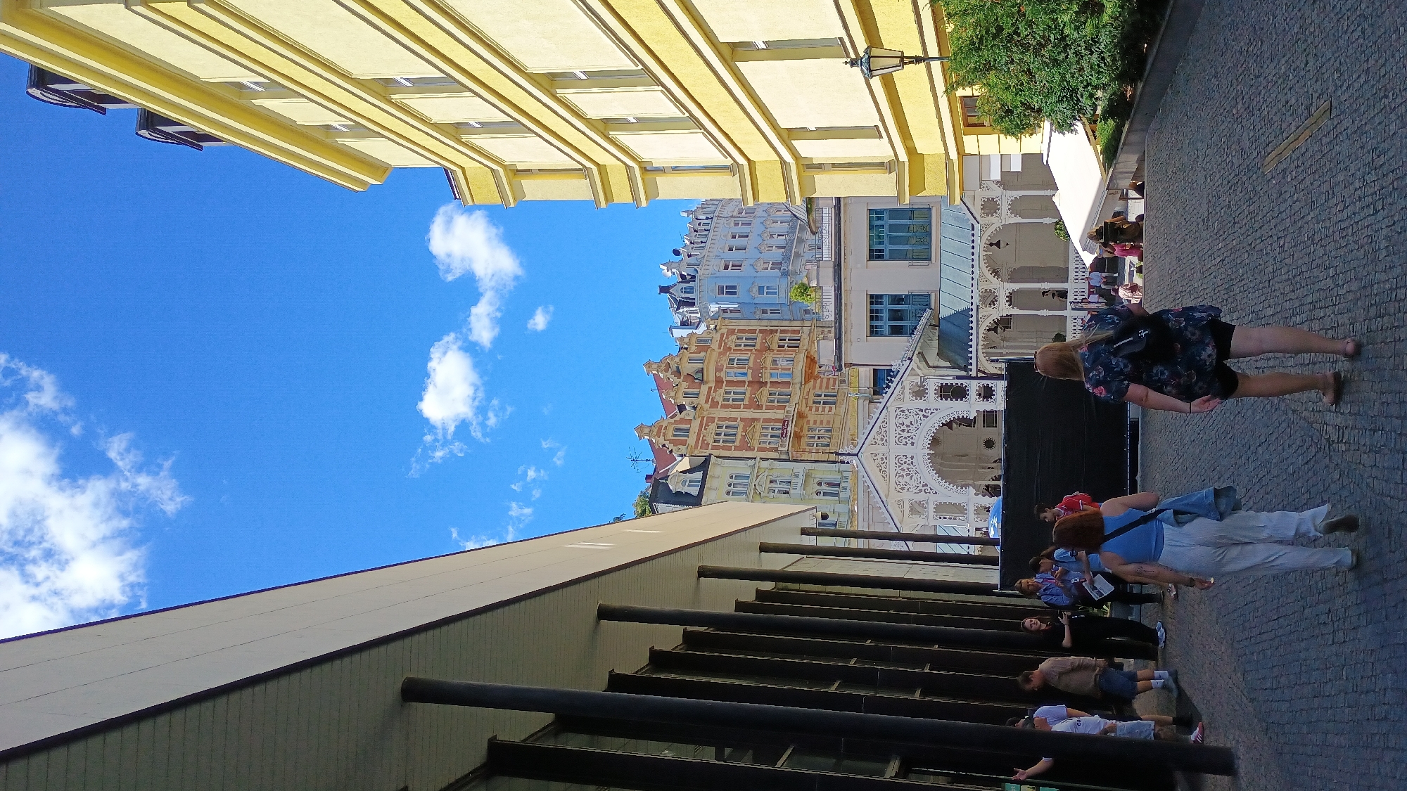 The image depicts a view of the colonnade in Karlovy Vary, Czechia, with the backdrop of colorful buildings. In the foreground, a paved walkway leads uphill. Two women are walking away from the camera on this walkway, one in a floral print dress and black shorts, the other in a blue top and white pants. To the left, a covered walkway with dark pillars frames the scene, and several people are standing and talking under it. Behind the walkway, ornate white colonnades extend, a characteristic feature of Karlovy Vary's spa architecture. Beyond the colonnades, a row of historic buildings with varying architectural styles and colors, including yellow, orange, and blue, are visible against a bright blue sky with scattered white clouds. A lamppost stands to the right of the yellow building.