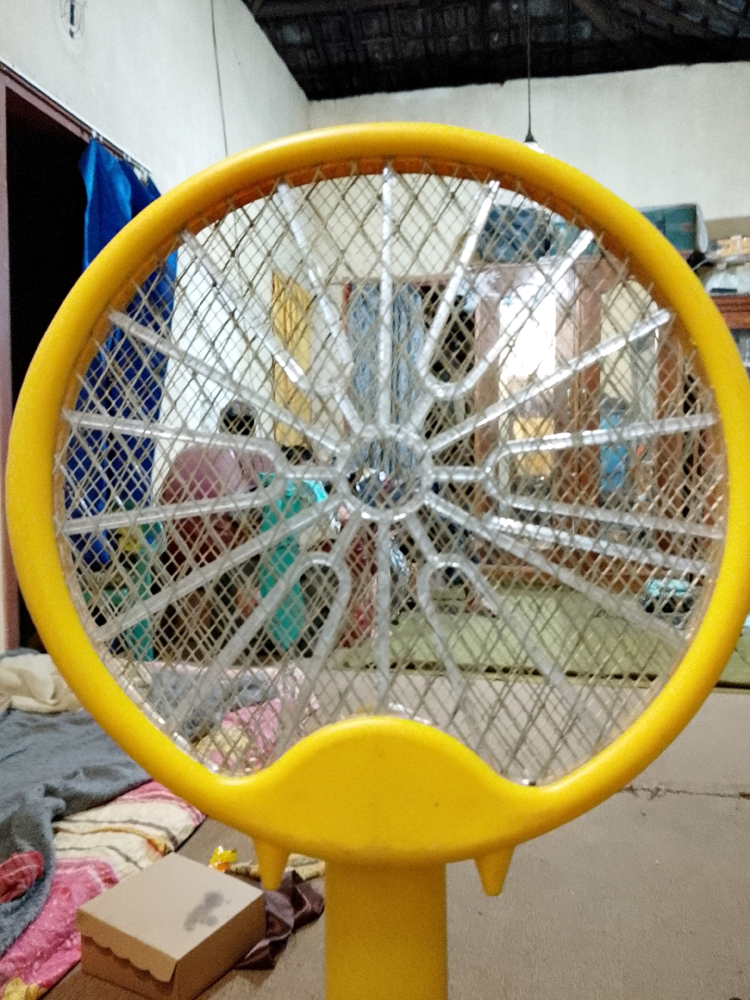 The image shows a close-up of a yellow electric mosquito racket with a metal mesh. The racket is positioned in the foreground, with its yellow frame and metal grid filling most of the frame. Behind the racket, a blurry background depicts an indoor scene. A person, appearing to be a child, is visible through the mesh, wearing a pink shirt and possibly a blue garment. Other indistinct figures and objects are also present in the background, suggesting a domestic environment. The floor appears to be made of concrete or a similar material. A cardboard box is visible in the lower left corner of the image, resting on a blanket. The overall lighting is dim. Based on the context provided by the prompt, this scene is located in Karangsari, Indonesia.