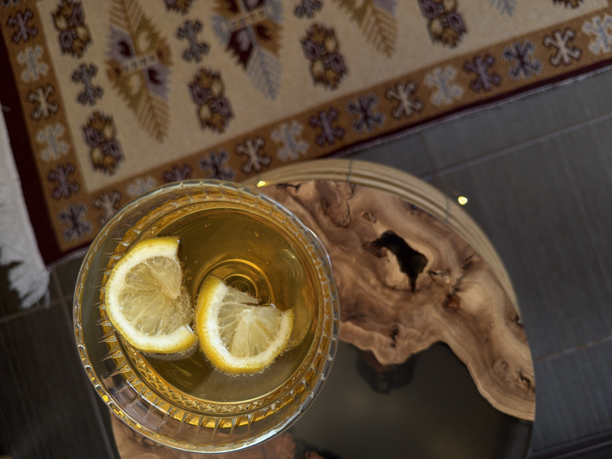 A high-angle shot shows a glass filled with a yellow liquid and two lemon slices, sitting on a round table with a wood grain design and a black glossy finish. In the background, a patterned rug in beige, brown, and burgundy tones partially covers the floor. The location is an interior space, presumably a home in an unknown city.
