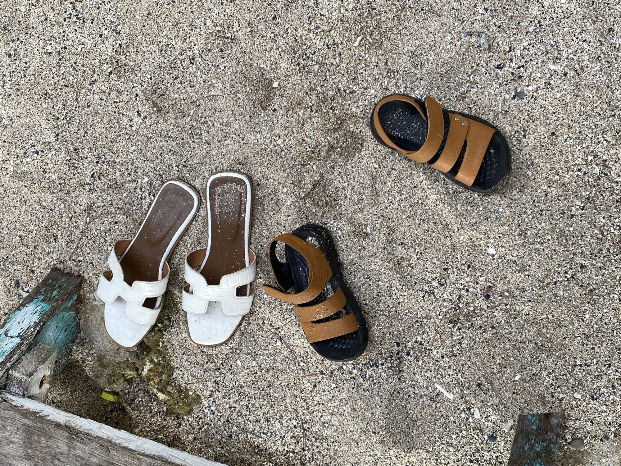 The image displays four pairs of sandals scattered on a sandy beach. Two pairs of white slip-on sandals with a distinctive H-shaped strap are positioned on the left side of the frame, partially near some weathered wooden debris and a small pool of water. On the right side, two pairs of brown and black sandals are visible. One pair is a simple slide sandal with a single strap across the toes, while the other is a more substantial sandal with multiple straps and a velcro closure. The sand is a mix of light-colored grains with scattered darker flecks and small shell fragments. The lighting suggests it is daytime, and the overall scene evokes a relaxed, casual atmosphere often associated with beach settings. There is no visible text in the image. The location is identified as Naga Umbang, Indonesia, which aligns with the sandy beach environment.