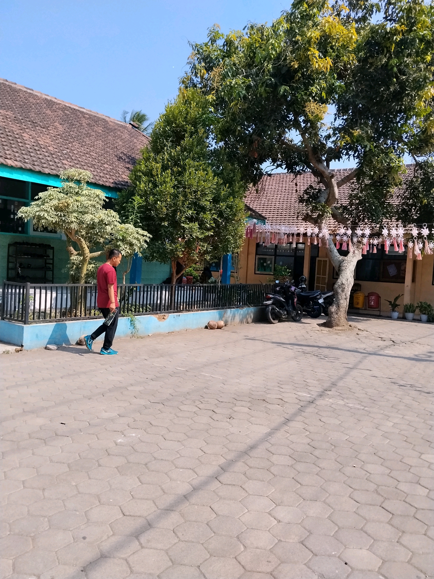 In Patemon, Indonesia, on a sunny day, a young boy wearing a maroon t-shirt and black pants is walking across a paved area in front of a school building. The school has a terracotta tile roof and a light blue trim. There are trees with green leaves and some yellowing foliage, and a large tree with a thick trunk is prominent in the foreground. The paved ground is made of interlocking hexagonal bricks. Two motorcycles are parked near the building. The scene is set outdoors, with a clear blue sky overhead.