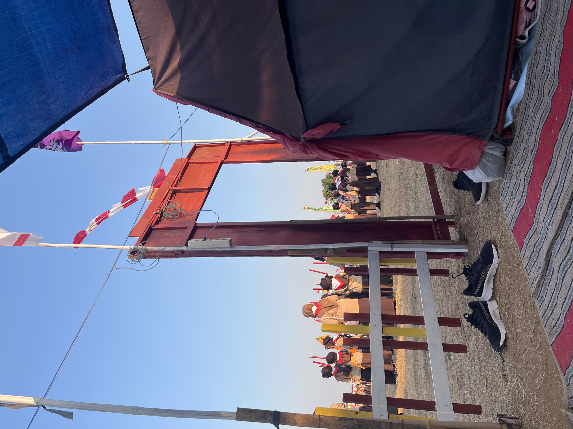The image depicts an outdoor scene under a clear blue sky, likely in Gembong, Indonesia, given the presence of scouts and Indonesian flags. In the foreground, a pair of black athletic shoes rests on the ground, adjacent to a colorful striped rug. A dark tent is pitched to the right, with a red fabric tie visible. Behind the tent and in the mid-ground, a red archway stands, with a blue tarp suspended above it. A group of individuals, appearing to be scouts in uniform with red neckerchiefs and headscarves, are gathered in the background, some holding long, red poles. Several flags are visible, including white and red striped flags and a purple flag with a symbol. The ground is covered in dry grass and dirt. A wooden fence, painted with white, red, and yellow stripes, separates the foreground from the area where the scouts are assembled.