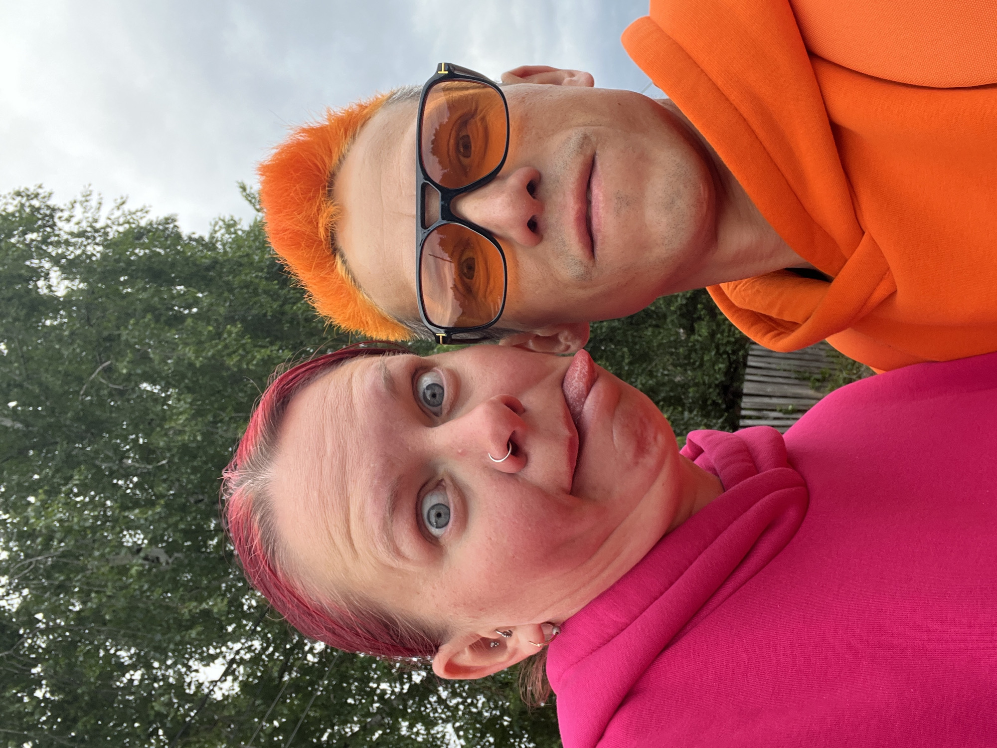 Two individuals pose for a selfie outdoors. The person on top has orange hair, wears sunglasses with amber-tinted lenses, and dons an orange hoodie. The person below has pink hair, a nose ring, is sticking their tongue out, and is wearing a pink hoodie. In the background, green trees are visible, partially obscuring a wooden fence. The photo was likely taken during the daytime under overcast conditions. The location is Chaikovsky, Russia.