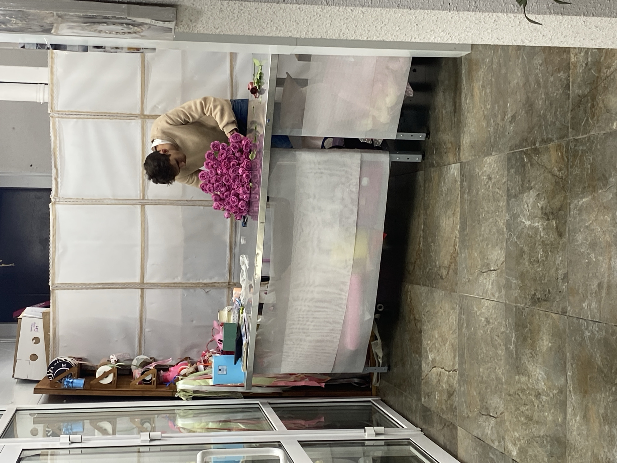 A person in a tan sweater stands at a counter, holding a bouquet of purple flowers. The counter is filled with various supplies, including boxes, ribbon rolls, and gift wrapping. Behind the counter is a white grid-like backdrop. The background suggests a retail environment, possibly a flower shop or gift shop. The floor is tiled with a stone-like pattern. There is a glass display case underneath the counter. It is unclear whether the location is in the city of Yekaterinburg, Russia.