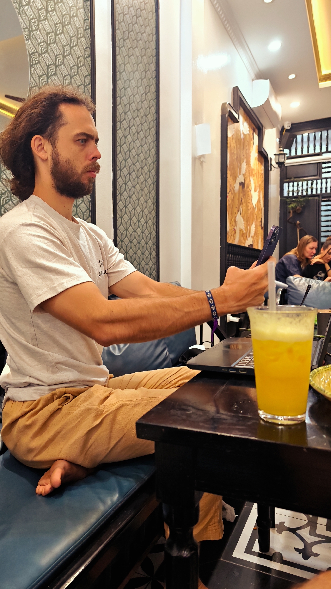 A man with medium-length curly hair and a beard is seated at a black table in what appears to be a cafe or restaurant in Phố Ràng, Vietnam. He is barefoot, wearing a light-colored t-shirt with faint text "ARGO" visible on it, and loose, light brown pants. His left arm rests on a dark blue cushioned bench, and he wears a blue and white patterned bracelet on his left wrist. He holds a purple smartphone horizontally in both hands, looking at it with a focused or serious expression. An open laptop is placed on the table in front of him. To his right, a glass of vibrant yellow, foamy drink with a straw is also on the table, alongside a small green bowl.
The setting features decorative wall panels with a patterned design on the left, and a white wall with recessed lighting and a wall lamp on the right. Further in the background, a large framed piece of artwork with golden tones hangs on the wall, and an air conditioning unit is visible above it. Beyond this, there is a dark wooden partition or window with a lattice design, and a decorative lantern. Two other individuals are seated in the blurred background, appearing relaxed. The environment suggests an indoor, well-lit space, likely a cafe where people are working or socializing.