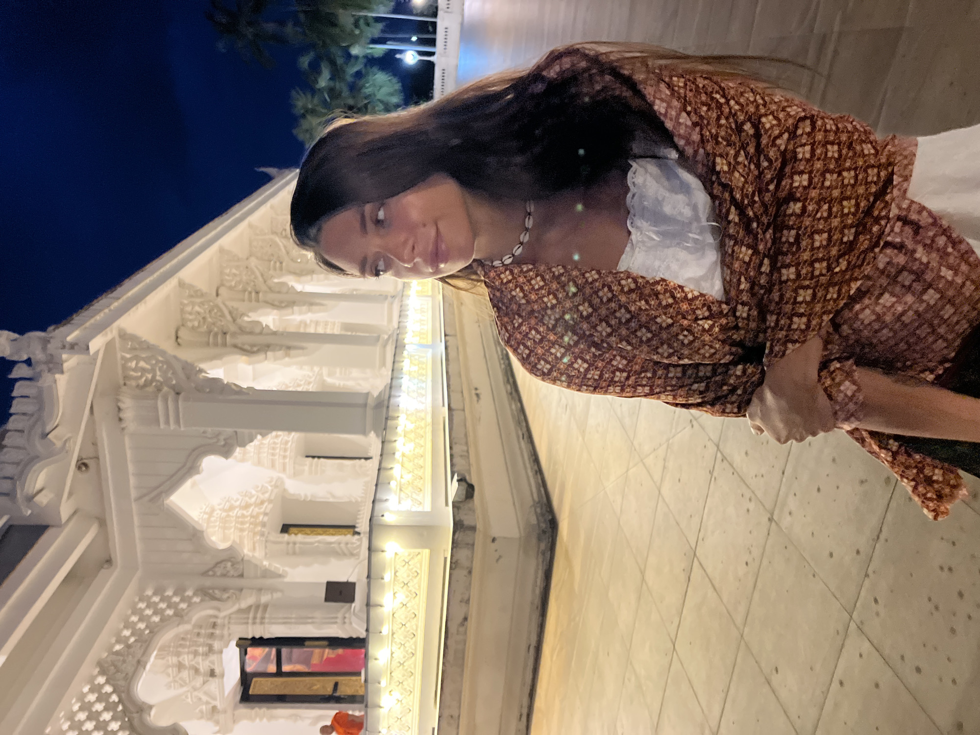 A woman stands in front of the illuminated Wat Plai Laem temple in Baan Bo Phut, Thailand. The temple's white facade is visible on the left side of the frame. The woman, facing slightly to the left, wears a light-colored top and a patterned brown and orange shawl. A seashell necklace adorns her neck. The ground beneath them is tiled. In the lower left corner, the blurred figure of a monk in orange robes is visible. The background sky is dark blue, with palm trees illuminated by spotlights.