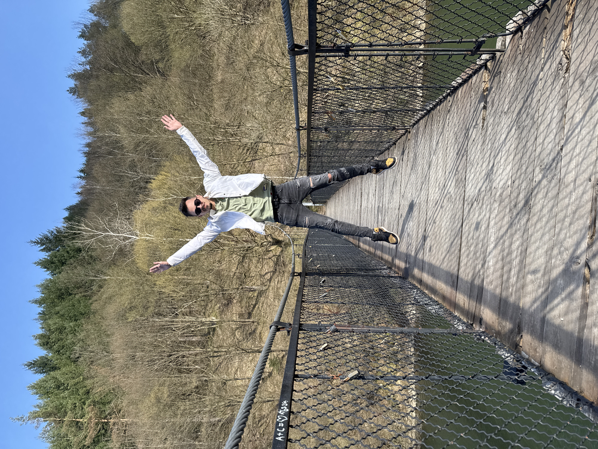 A man stands with outstretched arms on a narrow wooden bridge lined with metal chain-link fencing. He wears sunglasses, a light-colored jacket, a green shirt, dark ripped jeans, and sneakers. The bridge appears to cross a body of water. A forest covers the landscape in the background, under a clear blue sky. Graffiti is visible on the bridge support cables. The location is Dolni Pasarel, Bulgaria.