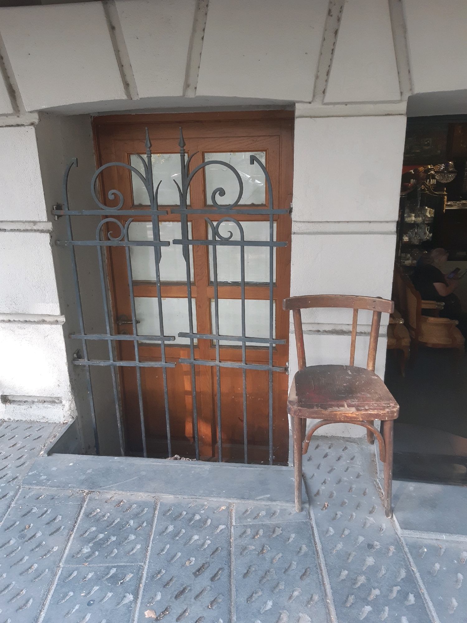 An old wooden chair with a worn seat and back rests on a textured metal surface outside a building in Tbilisi, Georgia. Behind the chair is a wooden door with glass panes, protected by an ornate metal grate. The building facade is light-colored with stone detailing above the door. To the right of the door, a glimpse into a dimly lit interior reveals shelves with various items and a person seated on another chair, looking at a phone. The metal surface in front of the building is divided into rectangular panels with a raised pattern.