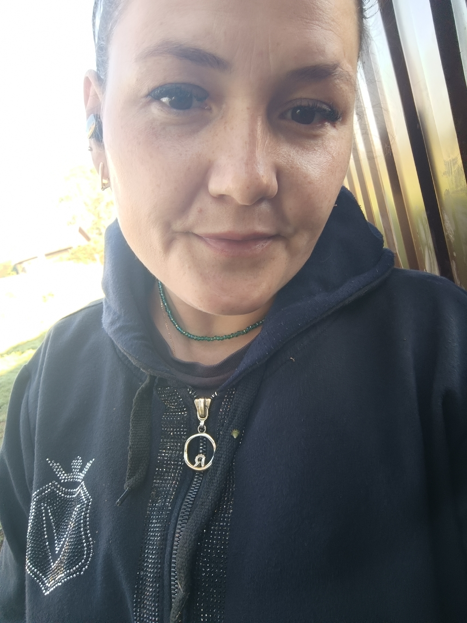 A close-up, medium shot captures a woman's face and upper body from the chest up, facing the camera. She has light brown skin with freckles and is wearing a dark blue hooded jacket. The jacket has a zipper and a decorative emblem on the left side, adorned with rhinestones. She is also wearing a green beaded necklace and a gold pendant with a circular design. Her hair is dark and pulled back, and she has a gold hoop earring in her visible left ear. In the background, a slatted wooden structure is visible, with hints of outdoor greenery and a bright sky. The scene appears to be outdoors.