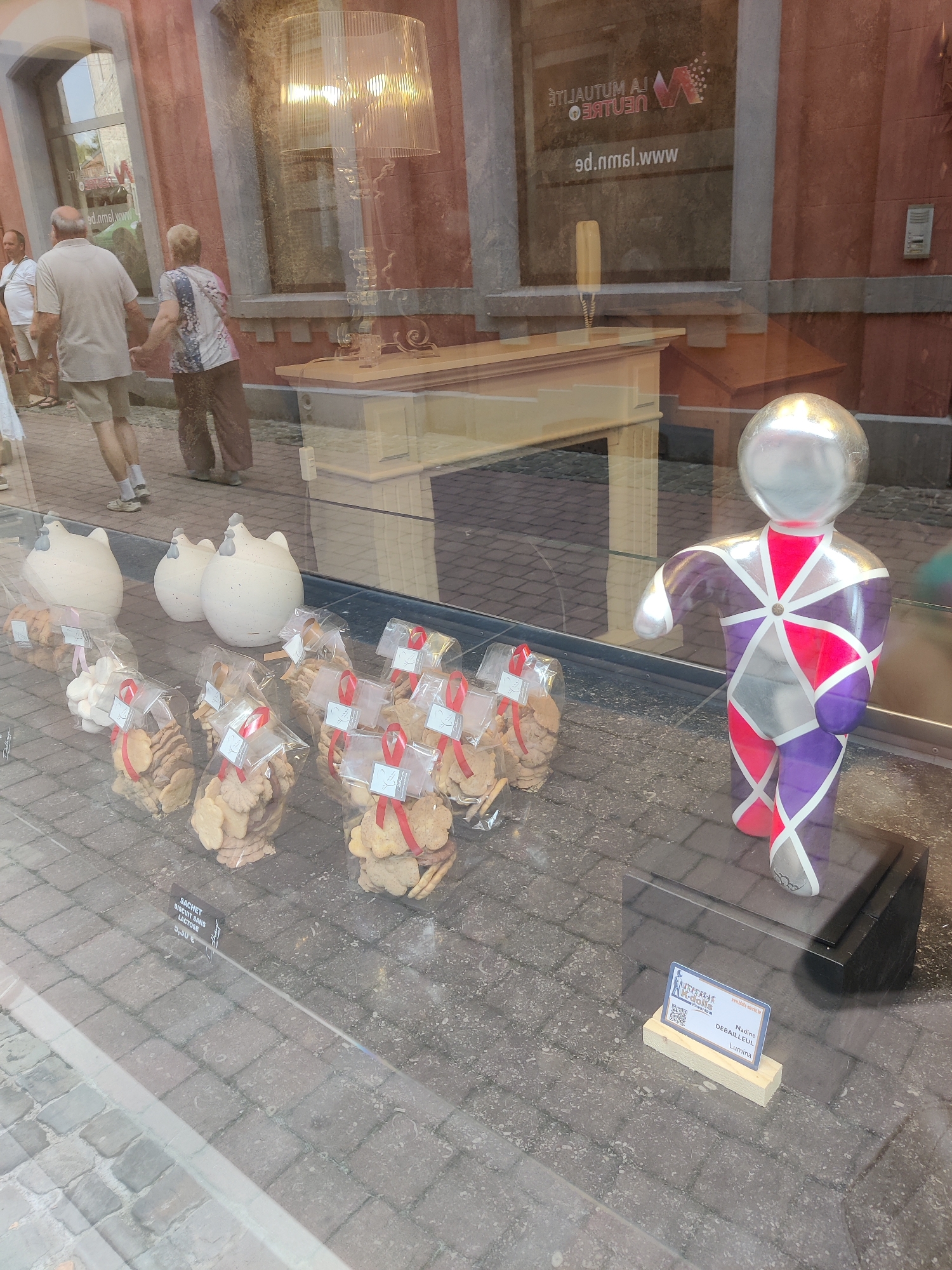 The image, taken through a shop window, shows a display of packaged cookies and decorative items on a cobblestone street. In the background, through the window, people are visible walking outside a building with a brick facade and large windows. A modern, large lamp is also visible inside the shop, as is a light-colored counter or display unit.
In the foreground, three white, stylized chicken-shaped ceramic figures are arranged on a display. To the right of the chicken figures, numerous clear plastic bags filled with cookies are displayed, each tied with a red ribbon and featuring a small white tag with a bird illustration. One of these bags has a label with French text, reading "SACHET BISCUITS SANS LACTOSE 3,30 €".
To the right of the cookies, a colorful, abstract sculpture of a person stands on a dark stand. The sculpture is predominantly silver, purple, and pink, with geometric patterns. In front of the sculpture, a small, light-colored wooden stand holds a blue and white business card. The text on the card includes "NAOMIE DEBAILLEUL LUMINA" and a website address. The presence of "Marche-en-Famenne, Belgium" on the business card identifies the location of this display.