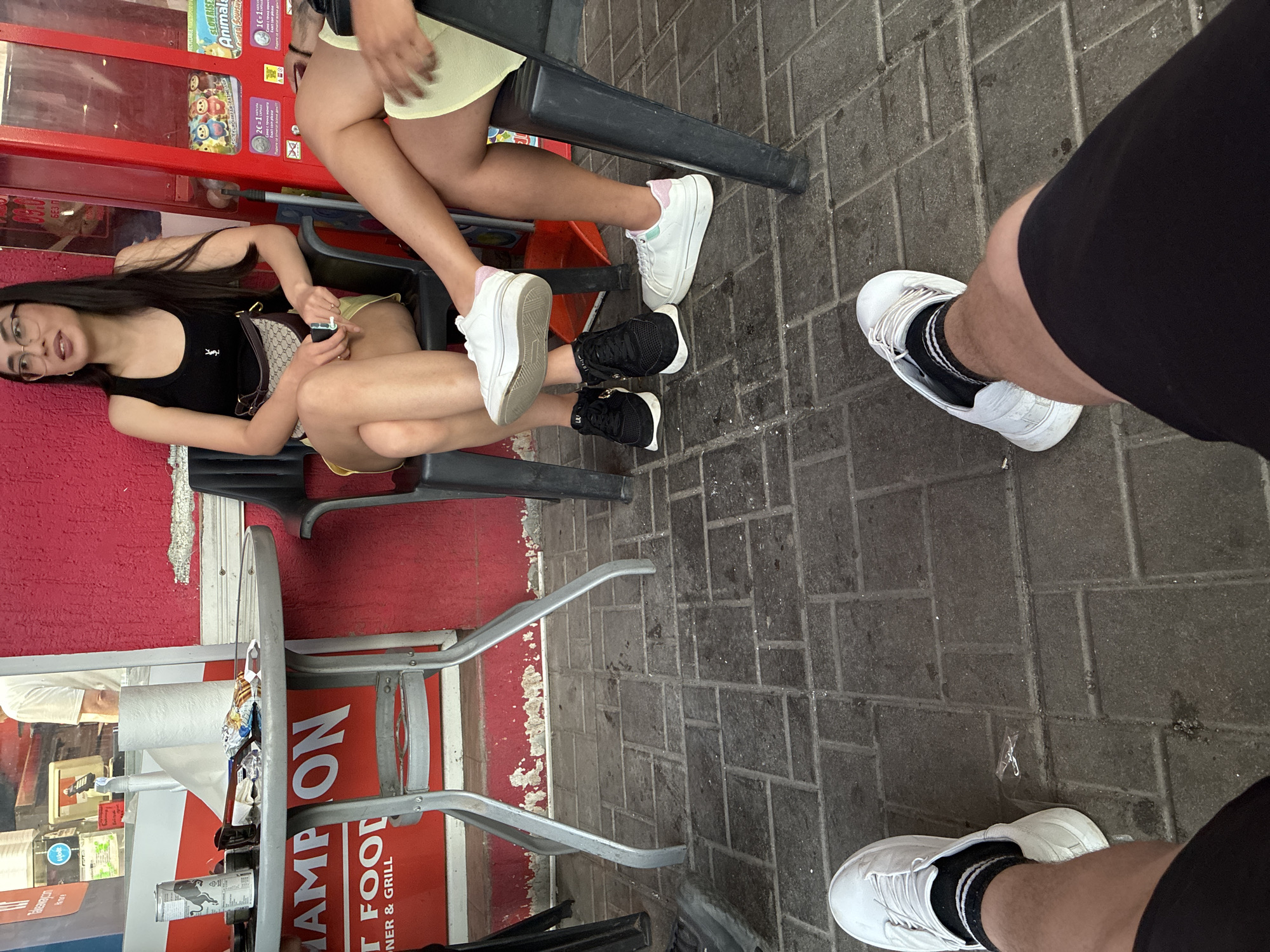 The image shows three people seated at a restaurant in Burgas, Bulgaria. Two women are sitting side-by-side at a table next to a red wall or structure with advertisements. One woman is wearing a black top and a light-colored skirt or shorts, and the other woman is wearing a light-colored top and yellow shorts. A third person, likely a man, is partially visible on the right side of the image, wearing black shorts and white sneakers. The table has a metal frame and is filled with various items, including what appears to be food packaging and a can. The ground is paved with gray bricks. The restaurant is identified as "Champion Food Diner & Grill" based on signage visible in the image.