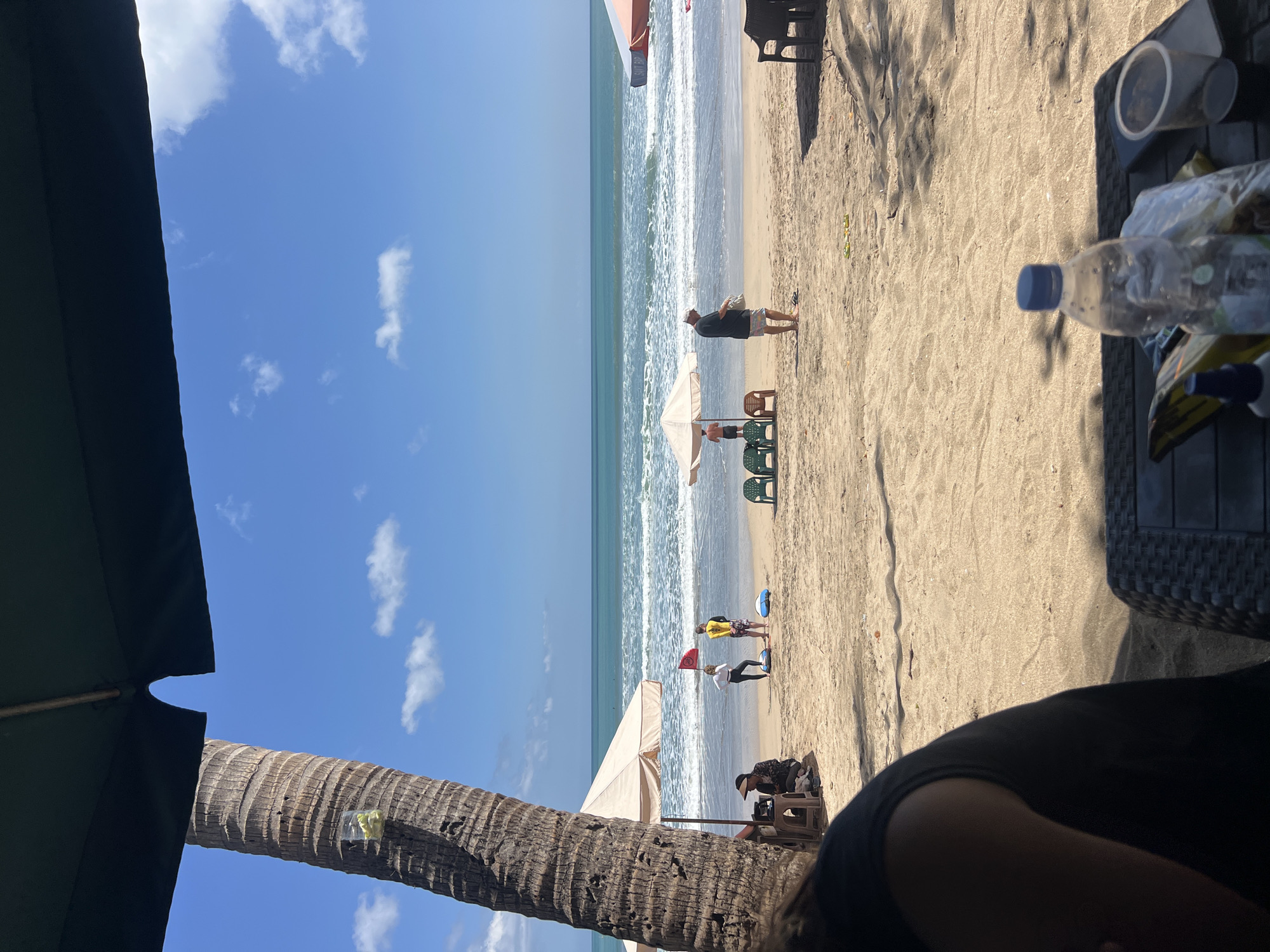 A sunny day at Kuta Beach, Bali, Indonesia. A thick palm tree trunk is on the left, with part of a dark green umbrella overhead. The sandy beach is populated with a few people, beach umbrellas, and chairs. In the foreground, a black table with a plastic water bottle and other items is visible. The ocean is calm with gentle waves breaking on the shore. The sky is clear blue with scattered white clouds.
