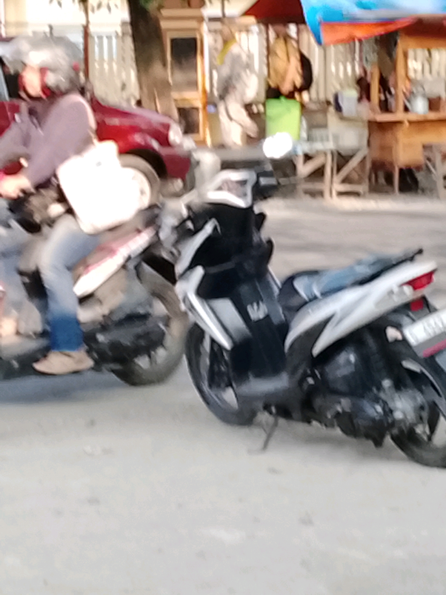 In Slawi, Indonesia, two scooters are visible on a paved road. On the left, a person wearing a helmet and a light-colored jacket is riding a scooter with a white bag attached to the front. This scooter is near a red car. To the right, a dark-colored scooter is parked. In the background, there are structures that appear to be market stalls with an orange and blue tarp overhead. The environment suggests a commercial area with people present.