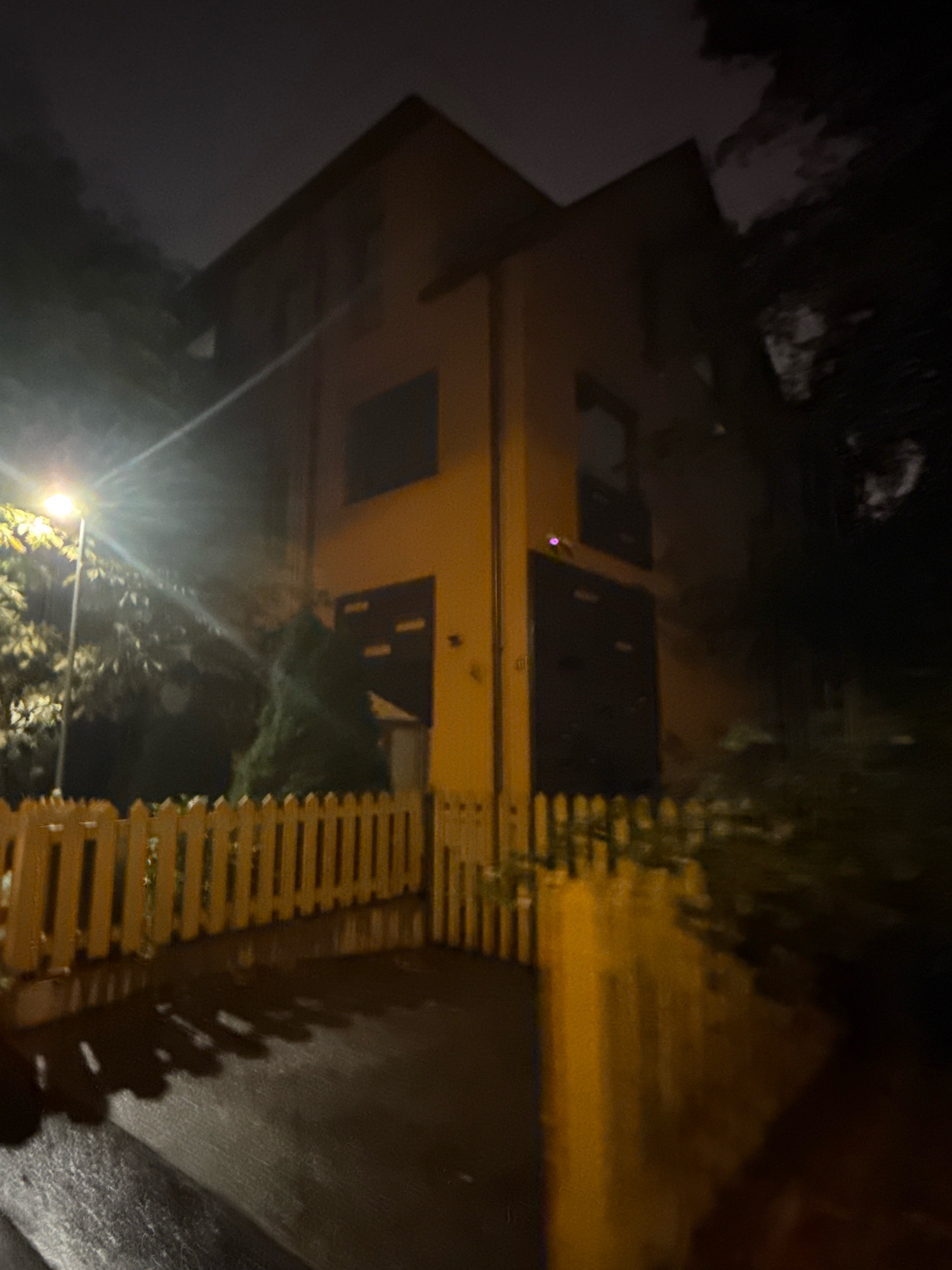 A dimly lit, grainy photograph taken at night shows a three-story house with a yellow facade and dark garage doors. A street lamp illuminates a portion of the house and the surrounding trees and vegetation. A white picket fence is visible in the foreground, with shadows cast on the wet pavement. The scene is set in Tallinn, Estonia.
