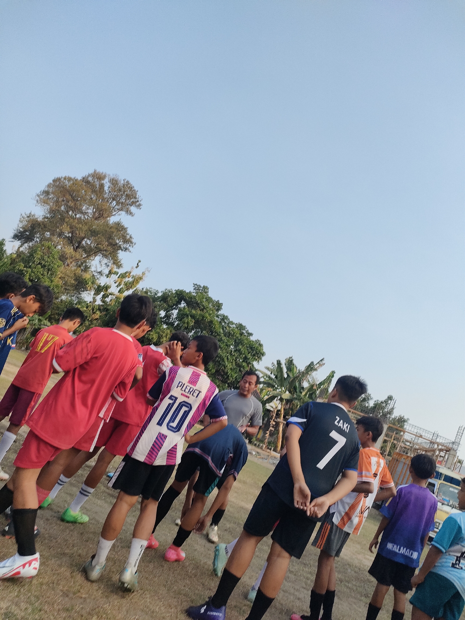 This is a low-angle, full-shot photograph taken outdoors on a grassy field. The scene depicts a group of young males, likely a soccer team, gathered together. Several individuals are wearing soccer uniforms with visible numbers and names on the back of their jerseys. One jersey has the name "PLERET" and the number "10". Another jersey has the name "ZAKI" and the number "7". A man, possibly a coach, is also present, wearing a grey shirt. The background features trees, foliage, and a clear blue sky. Some scaffolding and a portion of a white vehicle are visible in the distance on the right side of the image. The ground appears to be dry and composed of dirt and grass. The overall impression is a moment captured during a soccer practice or game in Banguntapan, Indonesia.