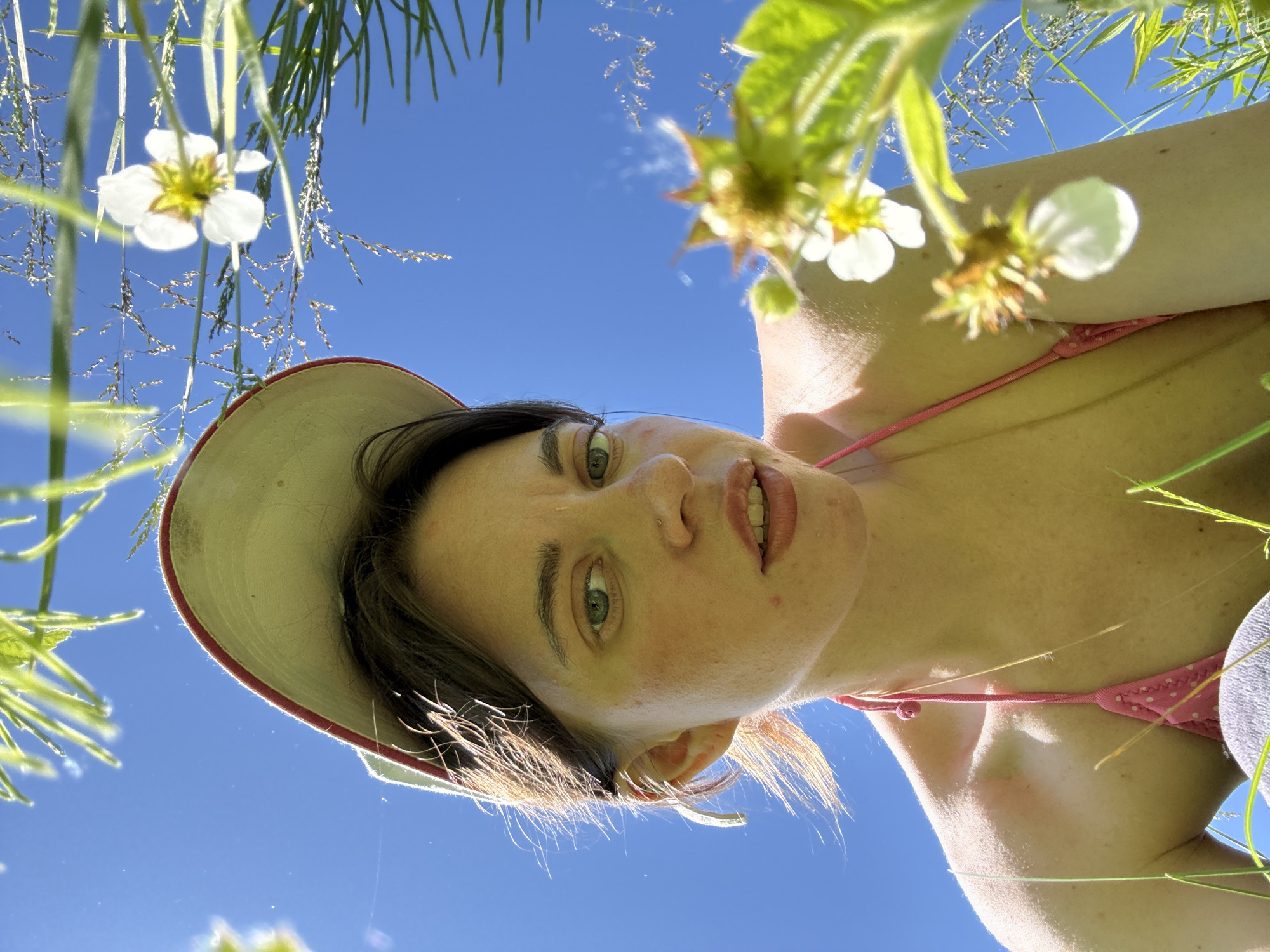 A low-angle shot shows a young Caucasian woman wearing a pink bikini and a red and tan baseball cap, looking upwards against a clear blue sky. She has light brown hair, green eyes, and light skin with freckles. She is surrounded by green grass and white flowers, some of which are in focus in the foreground. The lighting suggests it is a sunny day. The image was taken in Chaikovsky, Russia.