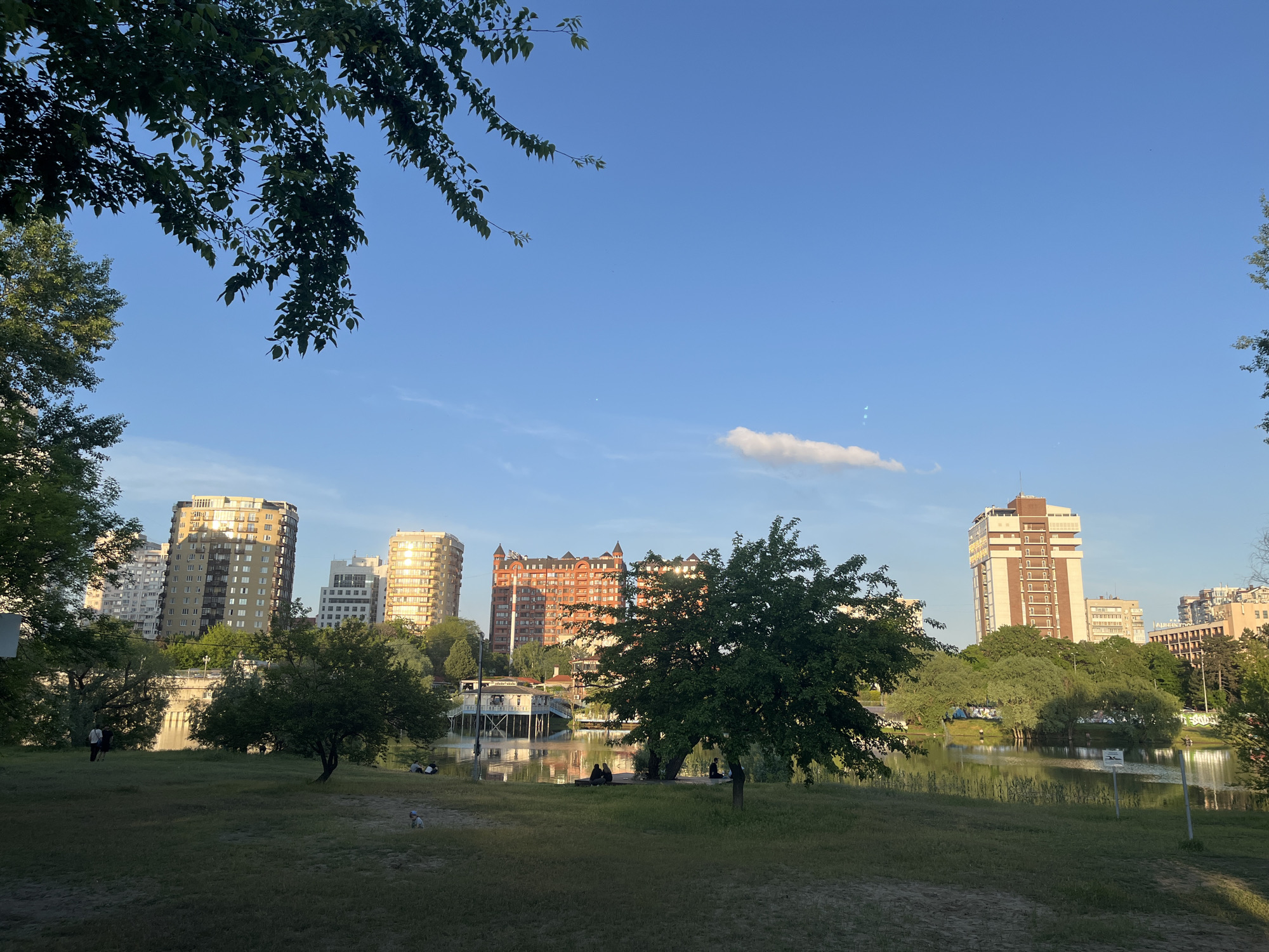 The image depicts a cityscape in Krasnodar, Russia, overlooking a body of water. Several high-rise buildings are visible in the background. The building in the center is red brick with some towers. In the foreground, there is a grassy area with trees and people relaxing near the water. A white cloud is in the blue sky.