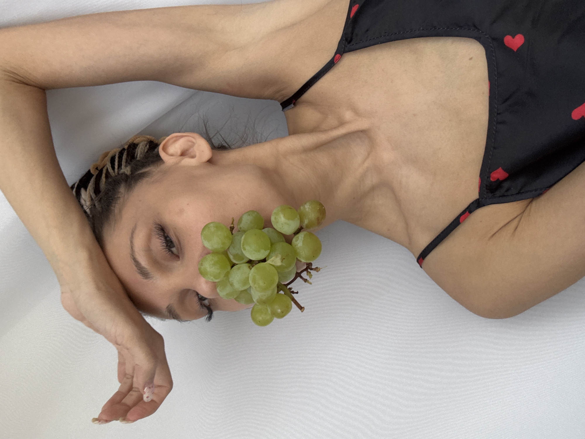 A woman lies on a white fabric, with a bunch of green grapes positioned in front of her mouth. She is wearing a black top with small red hearts. Her left arm is bent at the elbow and her hand is near her forehead. The location is unknown.