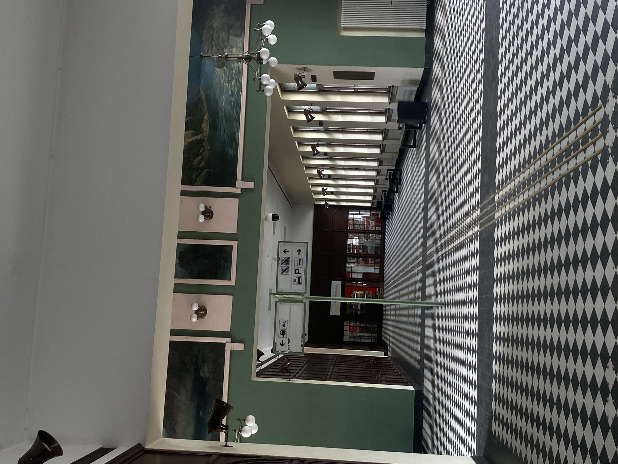The image depicts the interior of a train station hall in Nowy Sącz, Poland. The hall features a distinctive black and white checkered tile floor, which casts long, striped shadows from an unseen light source. On the left, a dark wooden wall with vertical panels is visible. Above, a green-painted wall displays framed landscape paintings. A chandelier with multiple lights hangs from the ceiling.
In the center of the hall, two directional signs are mounted on a green pole. The left sign indicates "WC" with an arrow pointing left. The right sign shows symbols for a person, a ticket, and a bus, with arrows pointing right, indicating facilities or directions for these. Further down the hall, through an open doorway, a red-doored building with glass windows, possibly a shop or ticket office, is partially visible.
Along the right wall, a series of tall windows with dark frames allow light to enter, illuminating the floor. Benches are placed along the wall in front of the windows. The overall atmosphere is spacious and somewhat empty, with a strong emphasis on geometric patterns from the floor and the lighting.