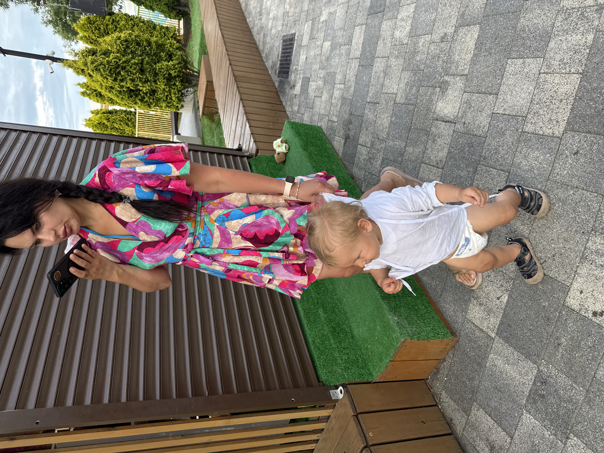 A woman with dark hair, styled in a braid, is standing and holding a smartphone to her ear. She is wearing a colorful, vibrant wrap dress. Beside her, a young child, likely a toddler, is standing and looking to the right. The child is wearing a white long-sleeved shirt and a diaper. The scene appears to be outdoors, on a paved area with a building with a brown corrugated metal exterior in the background. There are also some green bushes and wooden structures, suggesting a public space or park. The lighting suggests it is daytime. The location is the city of Sochi, Russia.