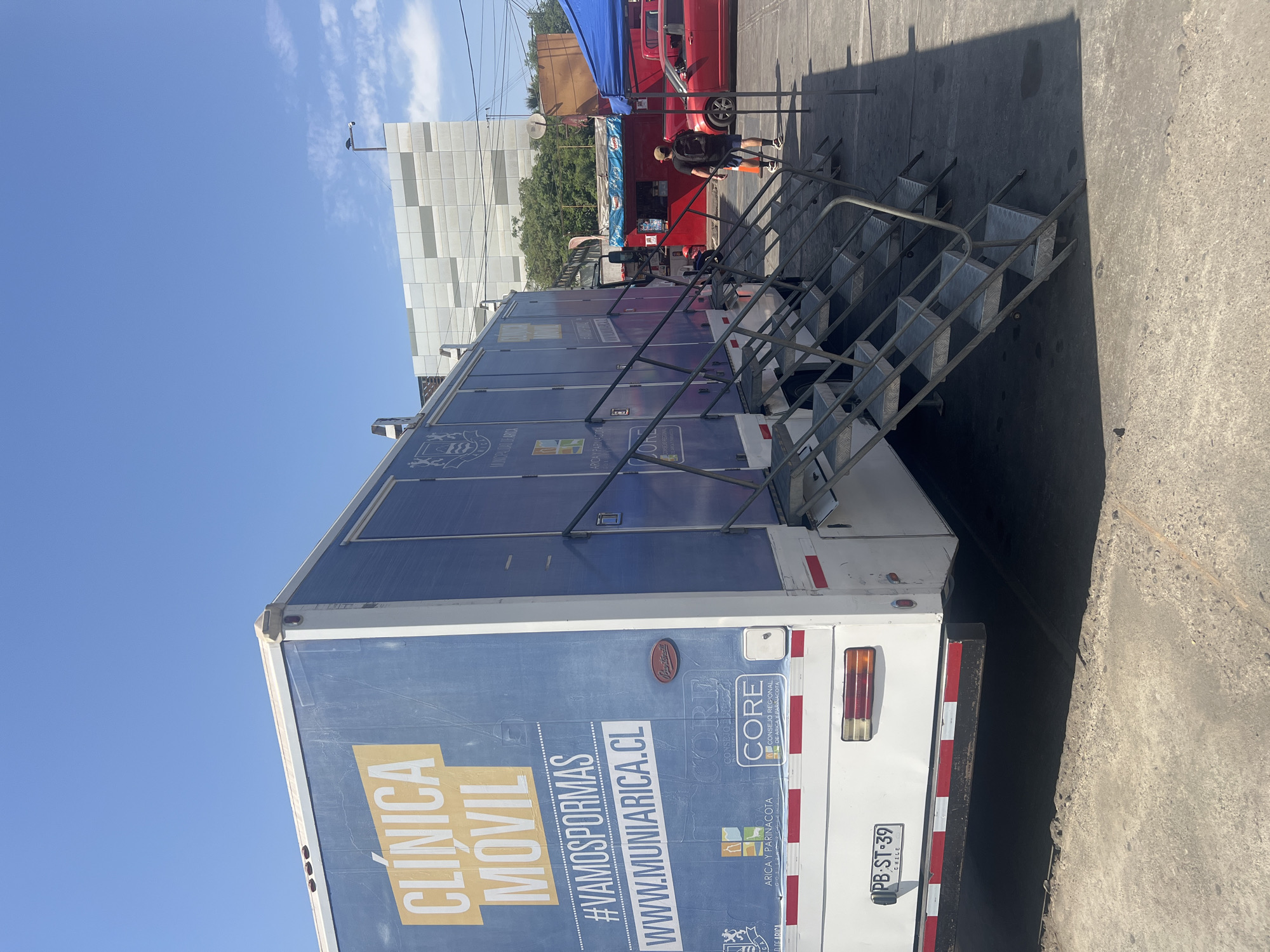 The image depicts a mobile clinic in Arica, Chile, as evidenced by the website address "WWW.MUNIARICA.CL" displayed on the side of the vehicle. The text "CLINICA MOVIL" is also prominent. The mobile clinic is white with blue graphics, and silver steps are attached to the side. A person is walking behind the mobile clinic. A red food truck sits behind the person. A building is visible in the background. The license plate of the mobile clinic is "PB-ST-39". The environment suggests an outdoor location within the city.