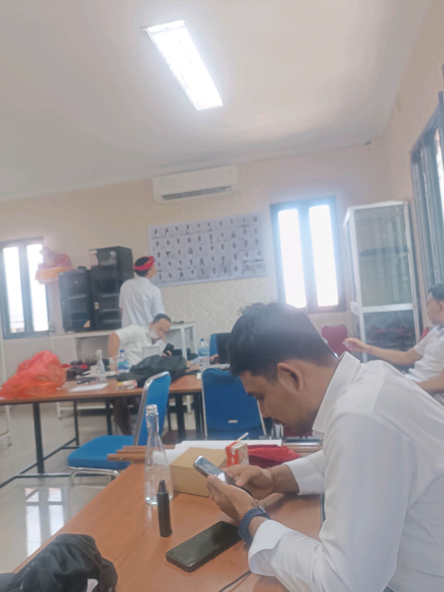 The image depicts an indoor scene with several individuals in what appears to be a classroom or meeting room. In the foreground, a man wearing a white shirt is seated at a wooden table, engrossed in his smartphone. Another person, also in a white shirt, is seated further back, looking down at a phone. To the left, another individual in a white shirt and red head covering stands near a set of speakers and a window. The room features a whiteboard with numerous small diagrams on the wall and large fluorescent ceiling lights. There are several tables and blue chairs, suggesting a collaborative or educational setting. Various objects are scattered on the tables, including bottles of water, a small cardboard box, and other personal belongings. The overall atmosphere suggests a moment of downtime or focus during a gathering. The specific location in Tabanan, Indonesia, cannot be determined from the visual information provided in the image.