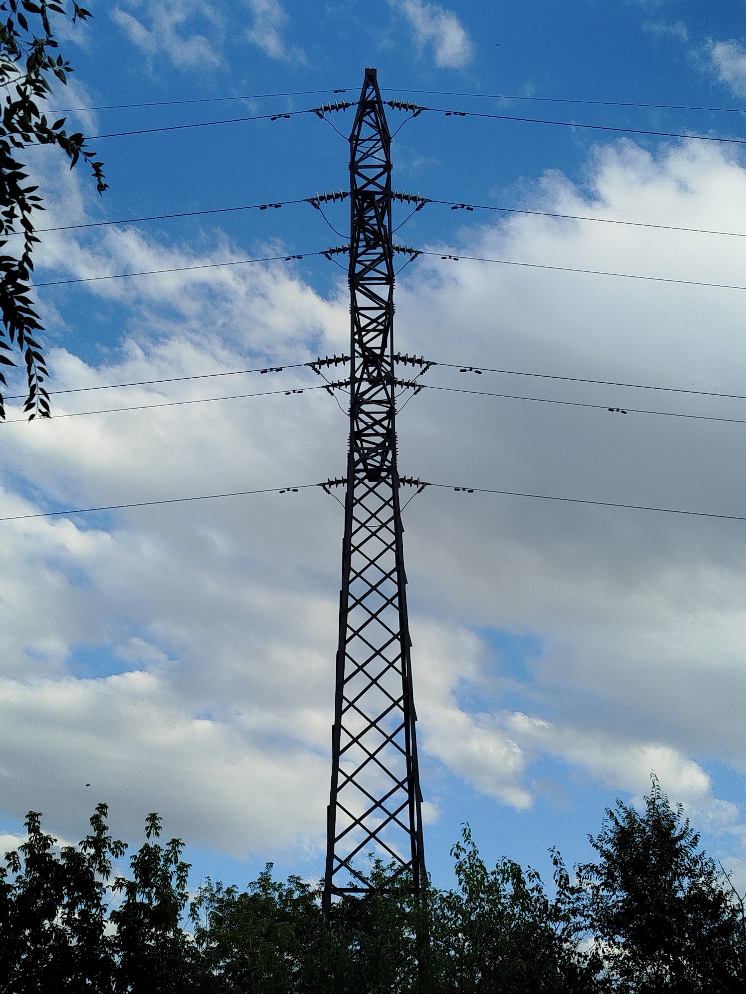 An outdoor, low-angle, vertical shot shows a dark metal power transmission tower against a bright blue sky with scattered white clouds. The tower is tall and lattice-like, with multiple horizontal crossbars supporting power lines. The lines stretch horizontally across the frame. In the foreground, silhouetted dark green trees and foliage are visible at the bottom of the image, partially obscuring the base of the tower. The overall scene suggests an industrial landscape integrated with nature. This image is presented without any specific identifiable landmarks that would pinpoint it to Qarağandı city administration, Kazakhstan.