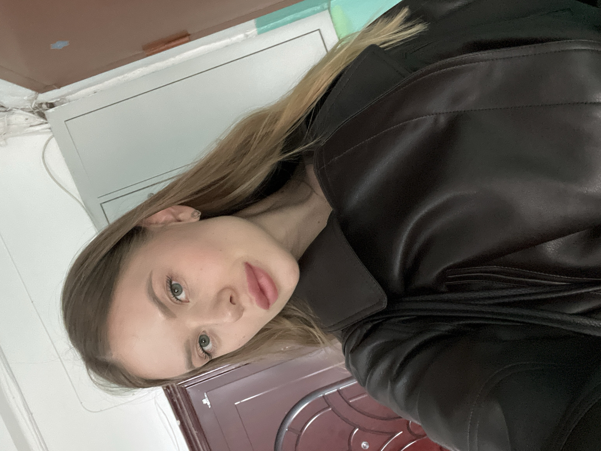 A young woman with long, straight blonde hair and light green eyes is captured in a close-up, rotated portrait, likely a selfie. She has light makeup, including pink lipstick. She is wearing a dark brown or black leather jacket with a structured collar and visible stitching details. Her expression is neutral and calm as she looks directly at the camera.
The background suggests an indoor setting, possibly within a home or apartment in Chaikovsky, Russia. To her left, there's a white wall. Above her, a light grey or white panel or cabinet is visible, with some exposed wiring or insulation near the top left corner where the wall meets a brown ceiling or fixture. To her right, a reddish-brown door with silver or white decorative patterns is partially visible. The lighting is soft, indicating an indoor environment where the time of day and weather are not discernible. No visible text is present in the image.