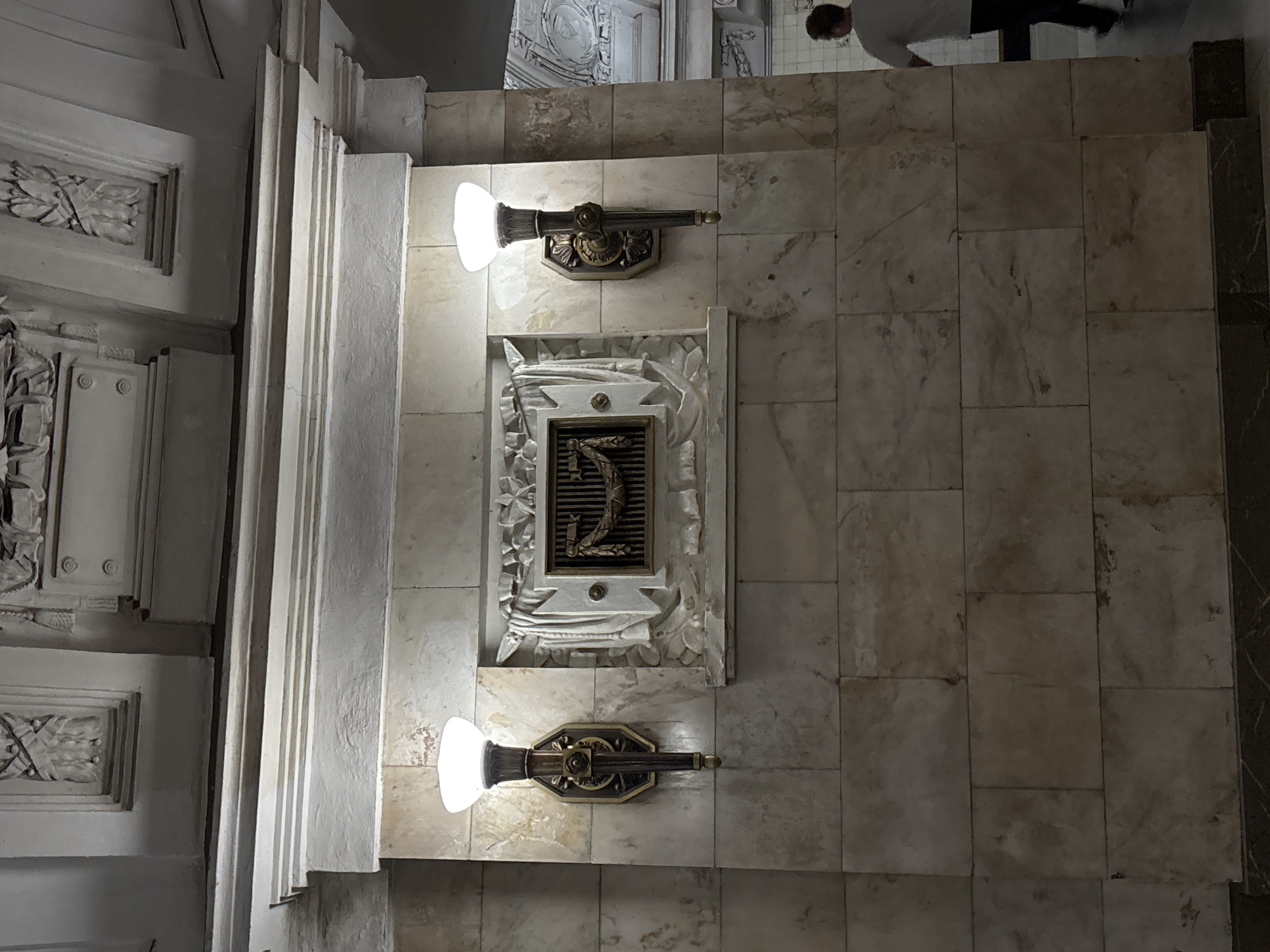 This is a photograph of a metro station interior in Moscow, Russia. The scene depicts a wall clad in light-colored marble with a decorative relief in the center. The relief is framed by a neoclassical architectural element and features a stylized garland. Flanking the relief are two ornate wall sconces, each topped with a frosted glass globe emitting a warm light. The ceiling above features intricate plasterwork and moldings. In the background, on the right side of the image, a person wearing a white t-shirt and dark pants is walking away from the camera. The overall ambiance suggests a grand and historical architectural style typical of some Moscow metro stations.