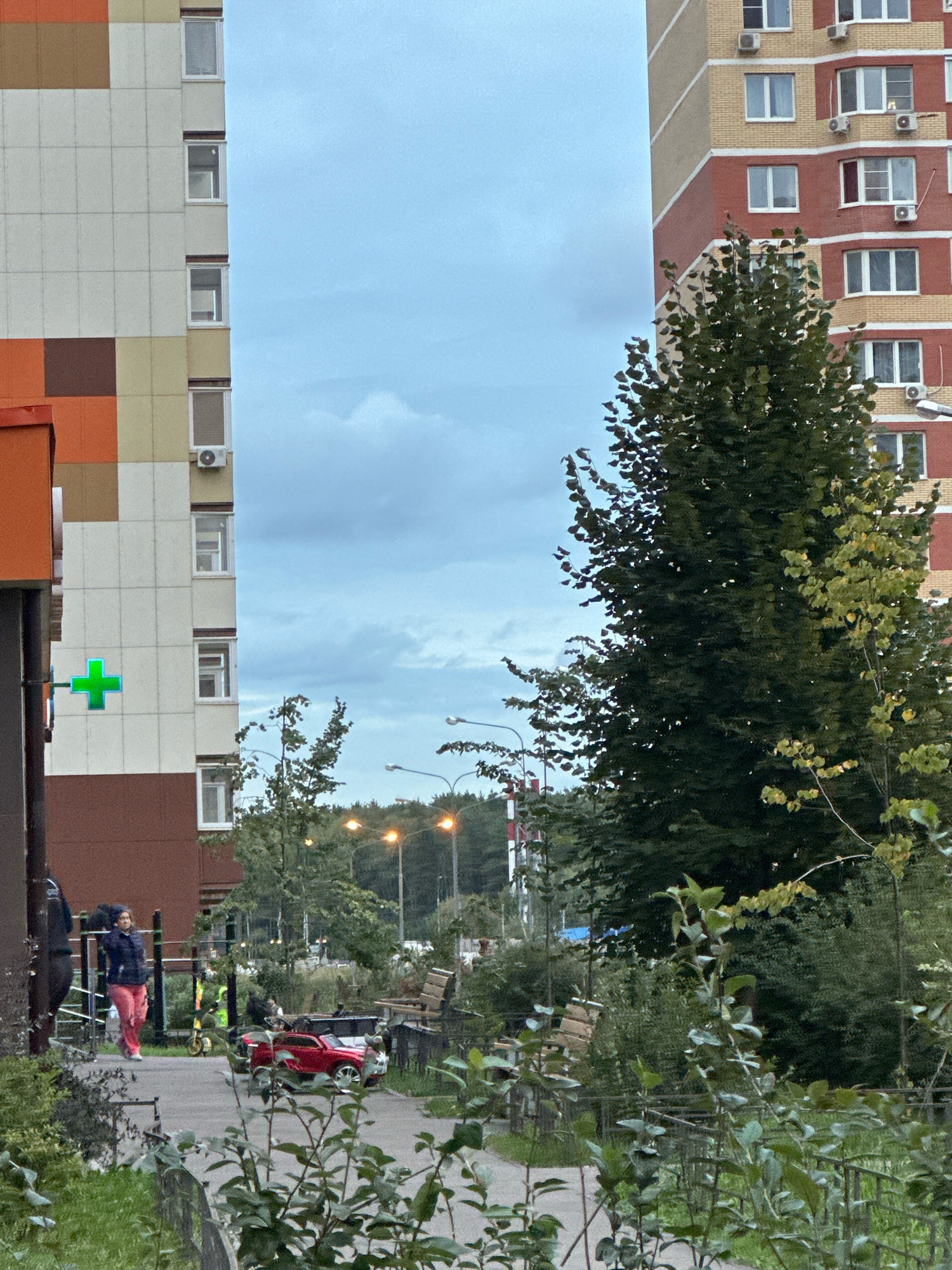 The image depicts a street scene in the city of Vidnoye, Russia. On the left, a multi-story building features a facade with alternating colored panels in shades of beige, white, brown, and orange. A bright green cross, likely indicating a pharmacy, is affixed to the building. To the right of this building is a gap, revealing a view of the sky and trees. Further to the right, another multi-story residential building with red and beige brickwork is visible, adorned with numerous air conditioning units. In the foreground, a paved walkway curves through an area with lush green bushes and trees. A woman in a dark jacket and pink pants is walking on the path. In the middle ground, a small red toy car is parked on the walkway. Streetlights and more trees are visible in the background. The sky is overcast with scattered clouds.