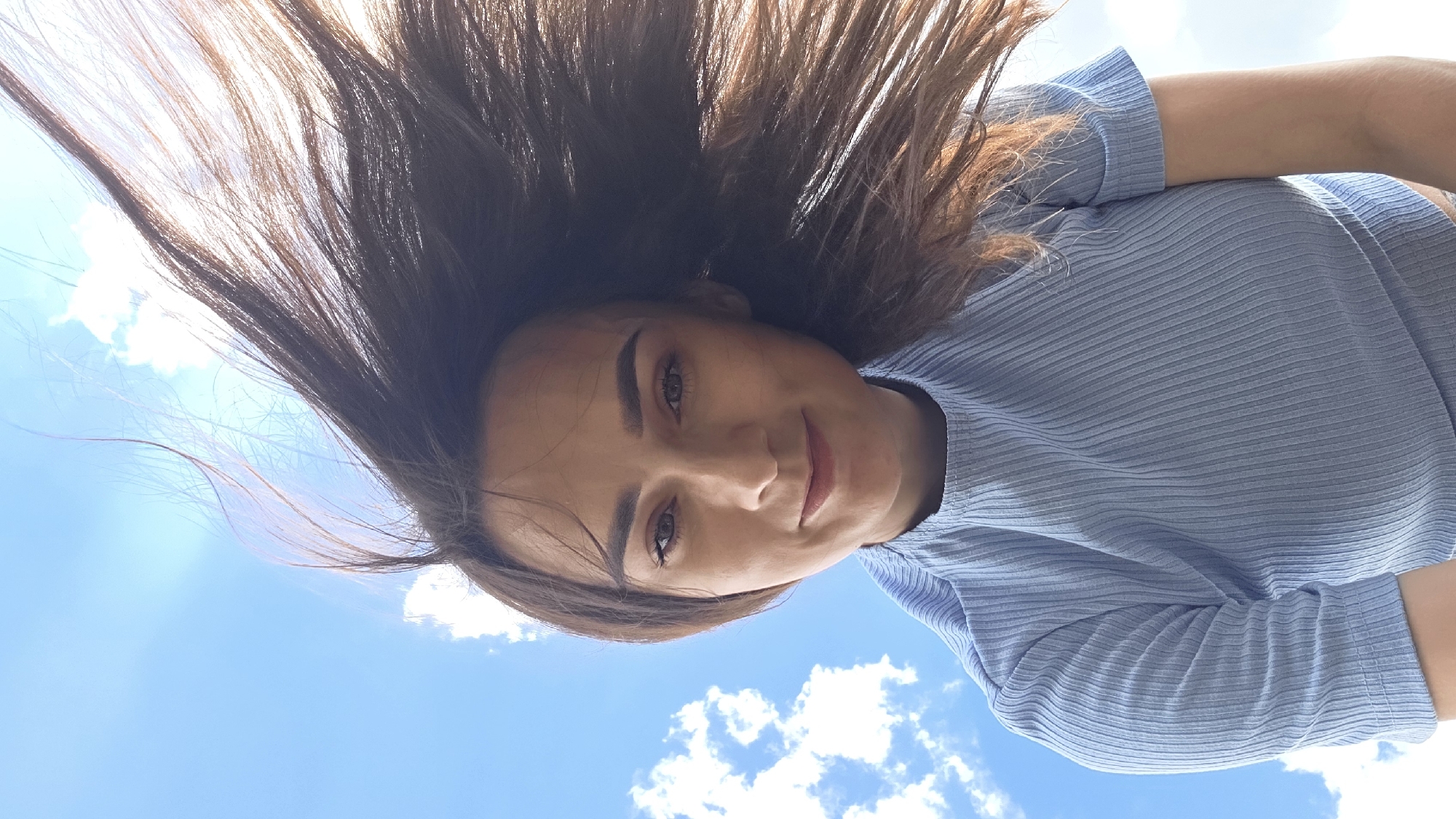 The media file is a close-up, upside-down selfie of a young woman with long, dark brown hair against a bright blue sky with scattered white clouds. She is wearing a light blue, ribbed, short-sleeved top. Her face is tilted upwards towards the camera, and she has a slight smile. Her eyes are brown, and she is wearing subtle makeup. Her hair is spread out, framing her face and suggesting movement, possibly from wind. The image appears to have been taken outdoors on a sunny day, likely during daylight hours. There are no visible text elements in the image. The setting appears to be an open outdoor space, with the sky being the dominant background element, indicative of a clear, pleasant day. The overall mood is relaxed and perhaps joyful.