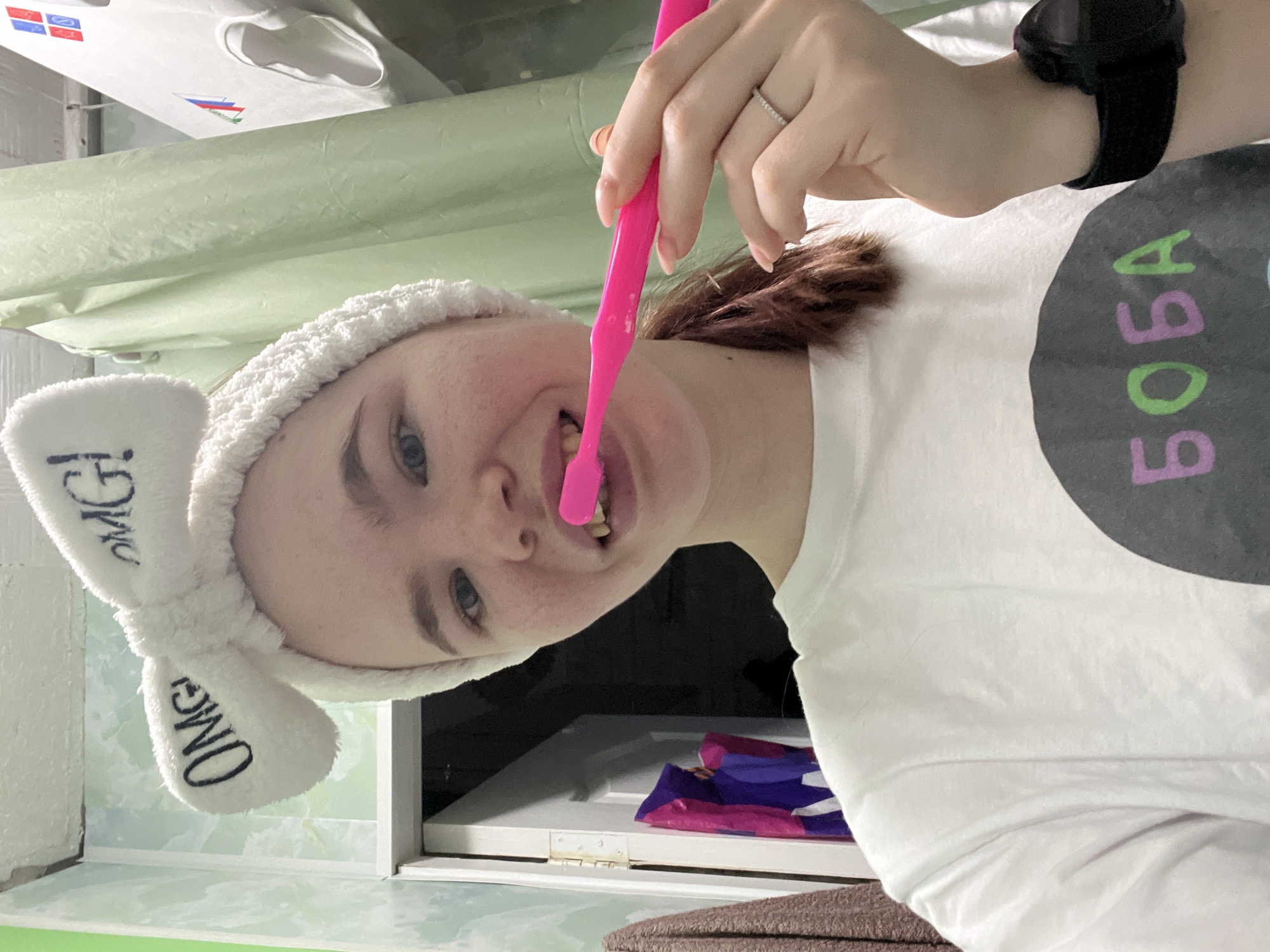 A young woman wearing a white headband with "OMG" printed on it and a white t-shirt with the word "BOBA" in black letters with green and purple accents is seen brushing her teeth with a pink toothbrush. She is wearing a black smartwatch on her left wrist and a ring on her ring finger. The scene appears to be set in a bathroom with a light green tile background. There is a white object with Russian flag colors visible in the upper left corner, possibly a container or piece of furniture. There are packaged sanitary products visible in the lower-center. The city of Chaikovsky, Russia is mentioned as the location of the scene.