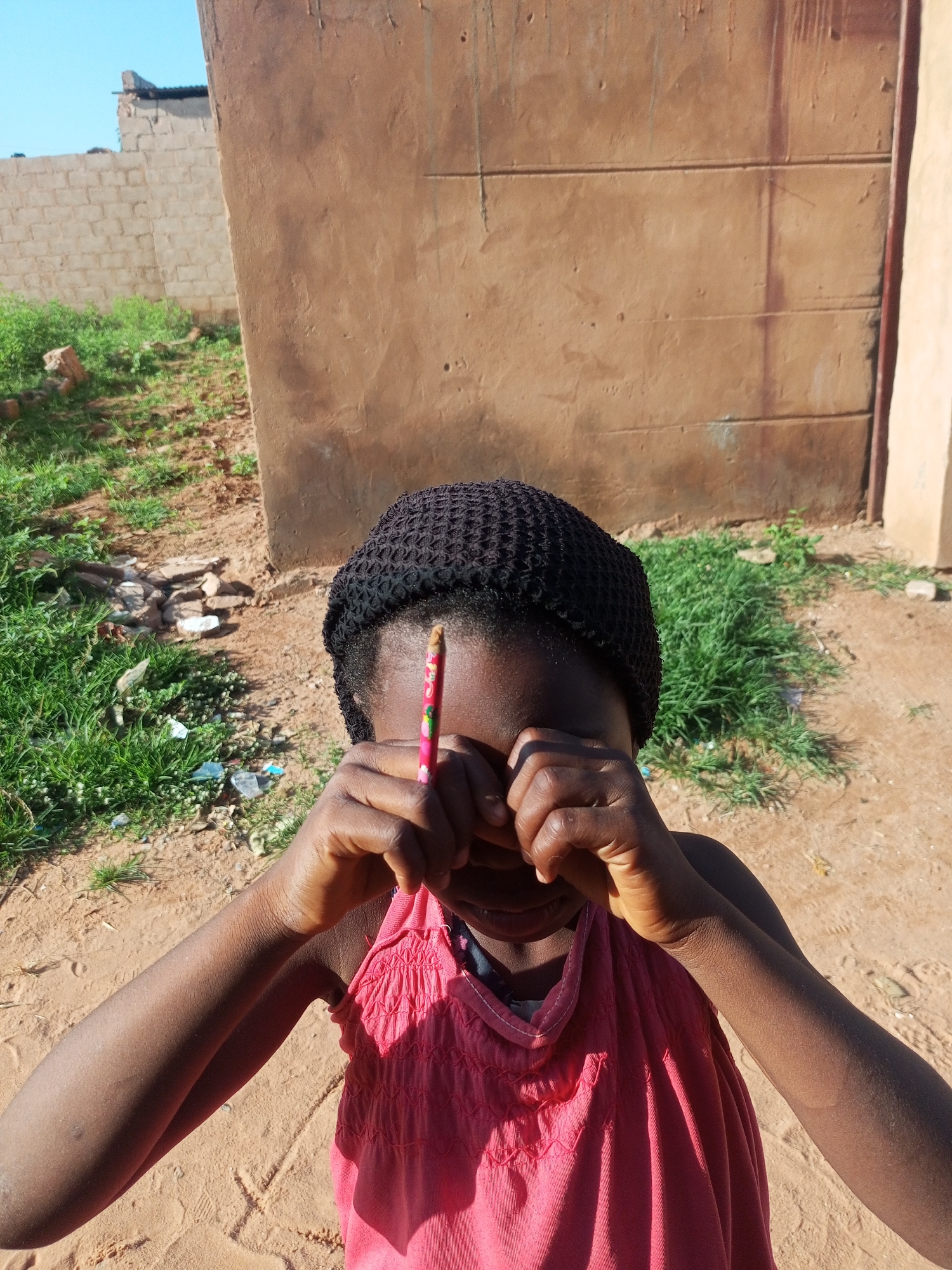 In Tempure, Nigeria, a young girl wearing a black head covering and a pink sleeveless top is positioned in the foreground, obscuring her face with her hands. A colored pencil is held between her fingers. The background features a rough, tan-colored wall with horizontal lines, and to the left, a patch of green grass with some scattered rocks and debris. The ground is sandy and dry. The sky is visible in the upper left corner, appearing light blue.