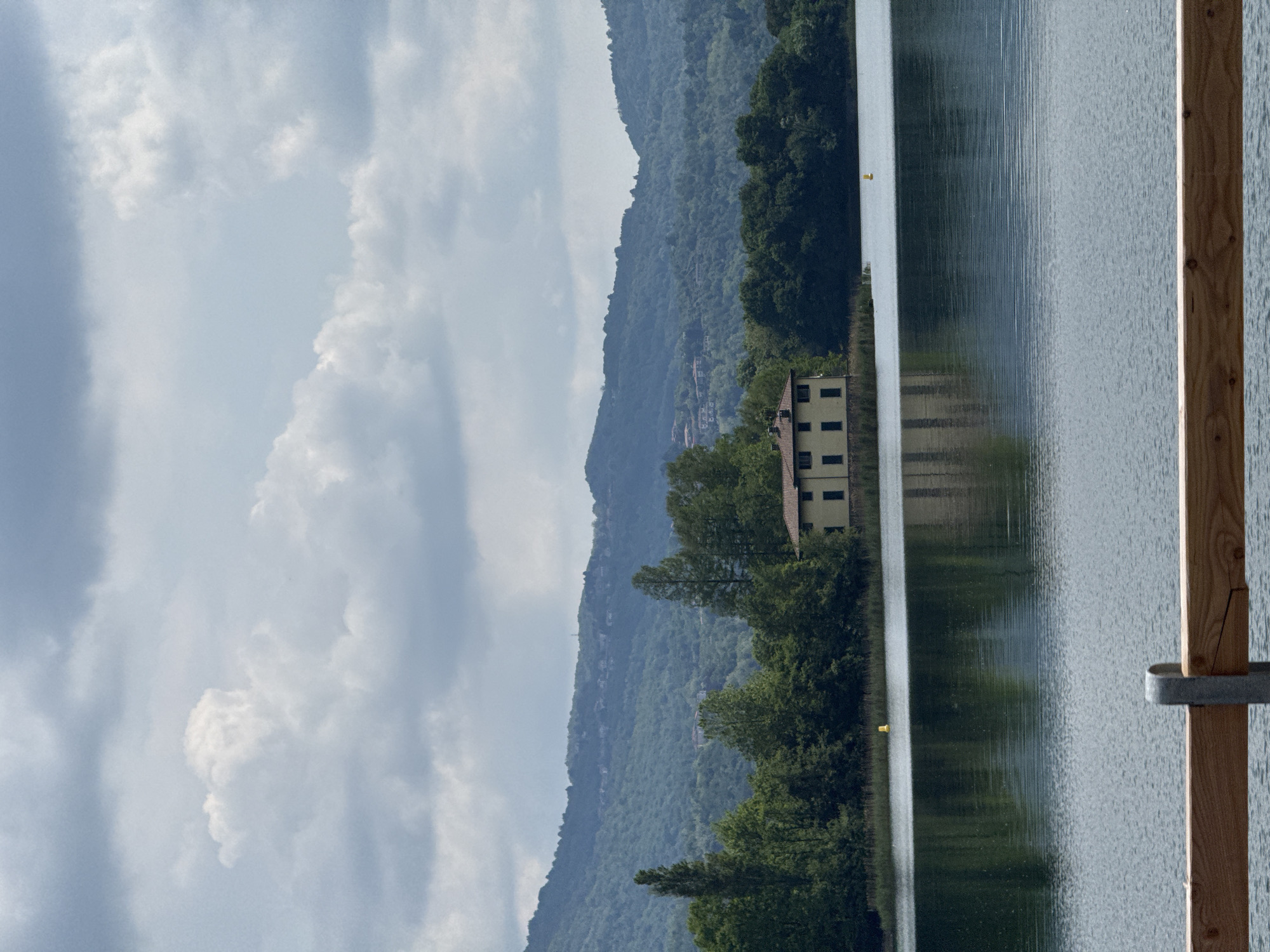 A tranquil landscape photograph captures a serene lake scene under a partly cloudy sky. The image, oriented horizontally, shows a body of water extending across the foreground and midground, reflecting the distant landscape and clouds. On the right side, a light-colored wooden post or railing, possibly part of a dock or pier, stands vertically, with a metal bracket attached. Further out on the water, a small yellow buoy-like object is visible.
Across the lake, the far shore is lined with lush green trees, partially obscuring a light yellow or beige multi-story building with multiple dark rectangular windows. Behind the trees and building, a range of rolling mountains or hills dominates the background, densely covered in verdant foliage. The sky above is a mix of light blue patches and large, fluffy white and gray clouds.
The scene appears to be set during daytime, likely late morning or early afternoon, given the bright yet diffused lighting. The weather is partly cloudy, contributing to the soft light on the landscape. The overall mood is peaceful and calm, with no specific activity taking place. There is no visible text in the image. The setting, located in Civate, Italy, strongly suggests the body of water is a lake, consistent with the region's geography.