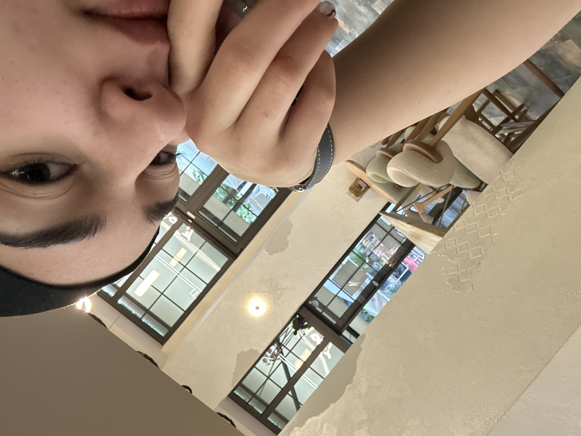 A first-person, upward-angled view shows a person's face, with their nose and left eye in focus, and their fingers touching their face. They are wearing a dark-colored head covering. Their wrist has a gray wristband. Background shows a window wall with visible daylight and seating chairs with beige-colored cushions. Based on the architecture, the location is likely a cafe or restaurant setting. The specific location and city, Burgas, Bulgaria, cannot be determined from the image alone.